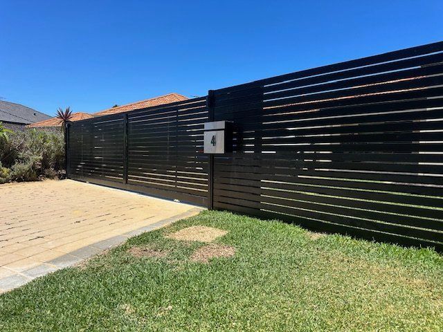 Black slatted fence with house number sign beside a paved driveway and green lawn under a blue sky