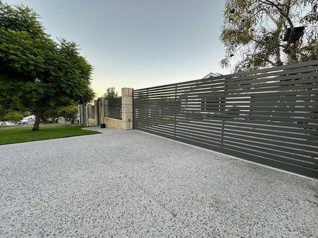 Gravel driveway beside a tall gray slatted fence, with trees and a lawn at left at dusk