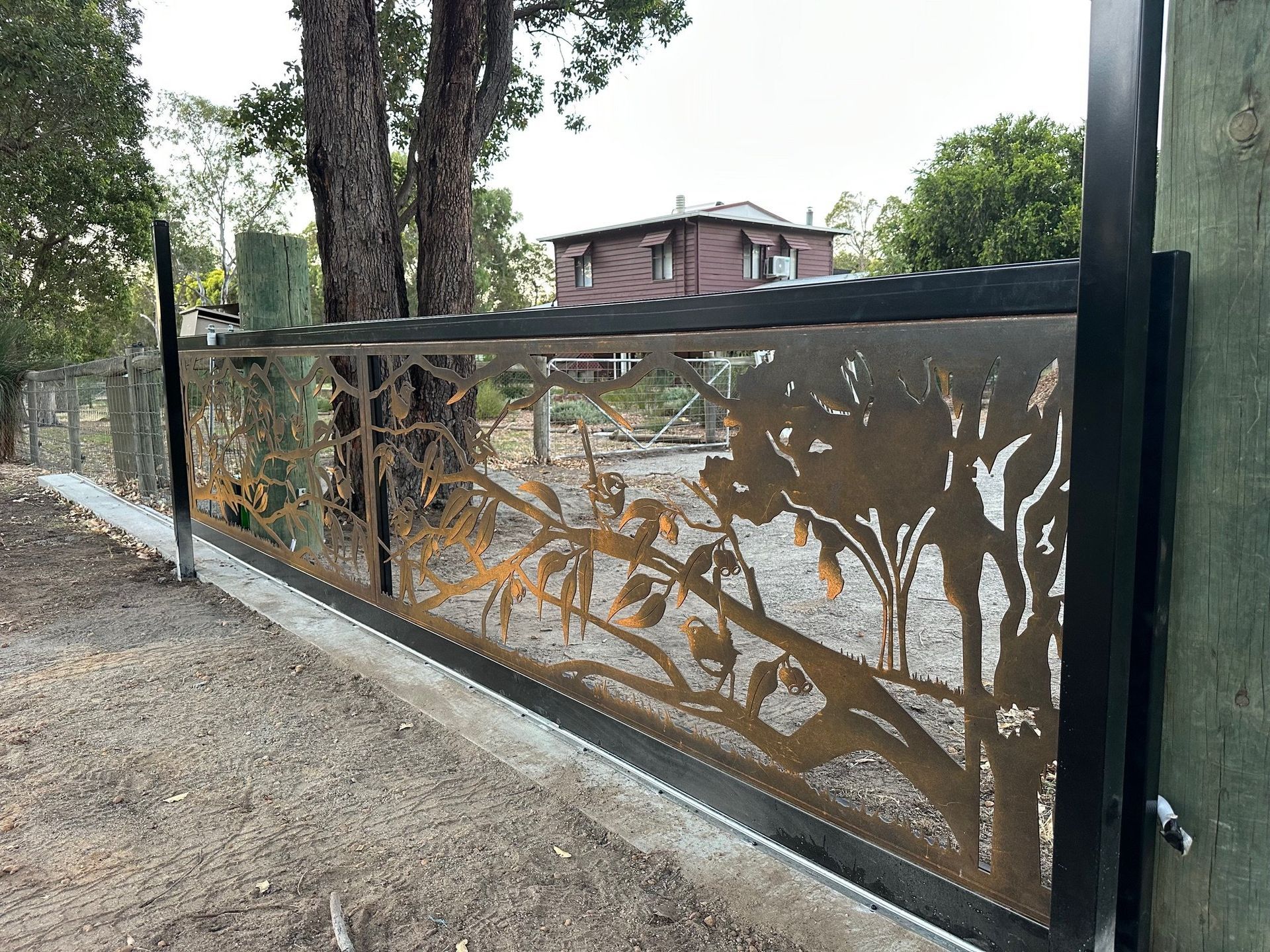 Rusty metal gate with tree and leaf cutout pattern beside a dirt path and trees outside a house
