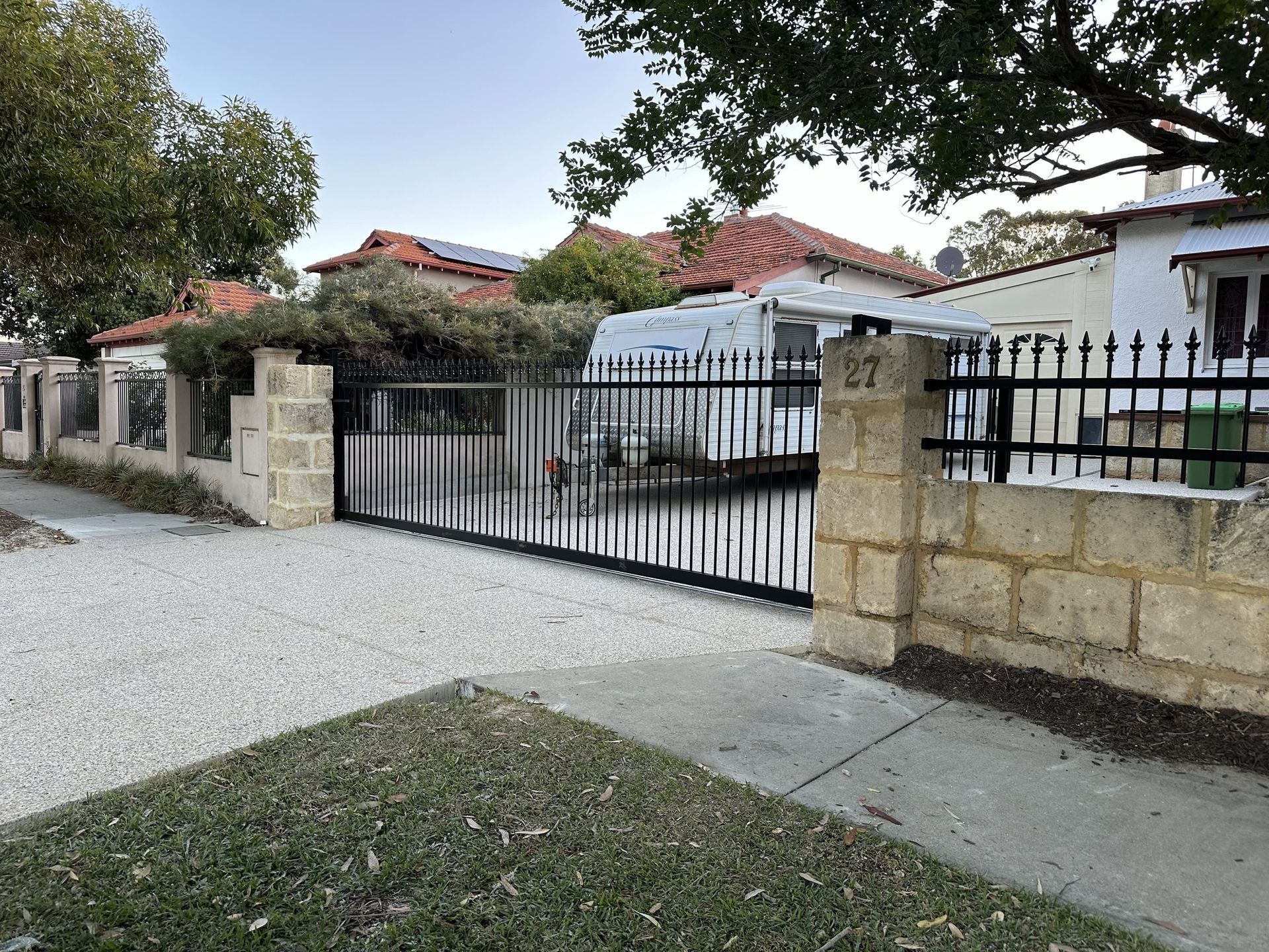 Residential driveway with black metal gate and stone wall, trees and red-roof houses behind.