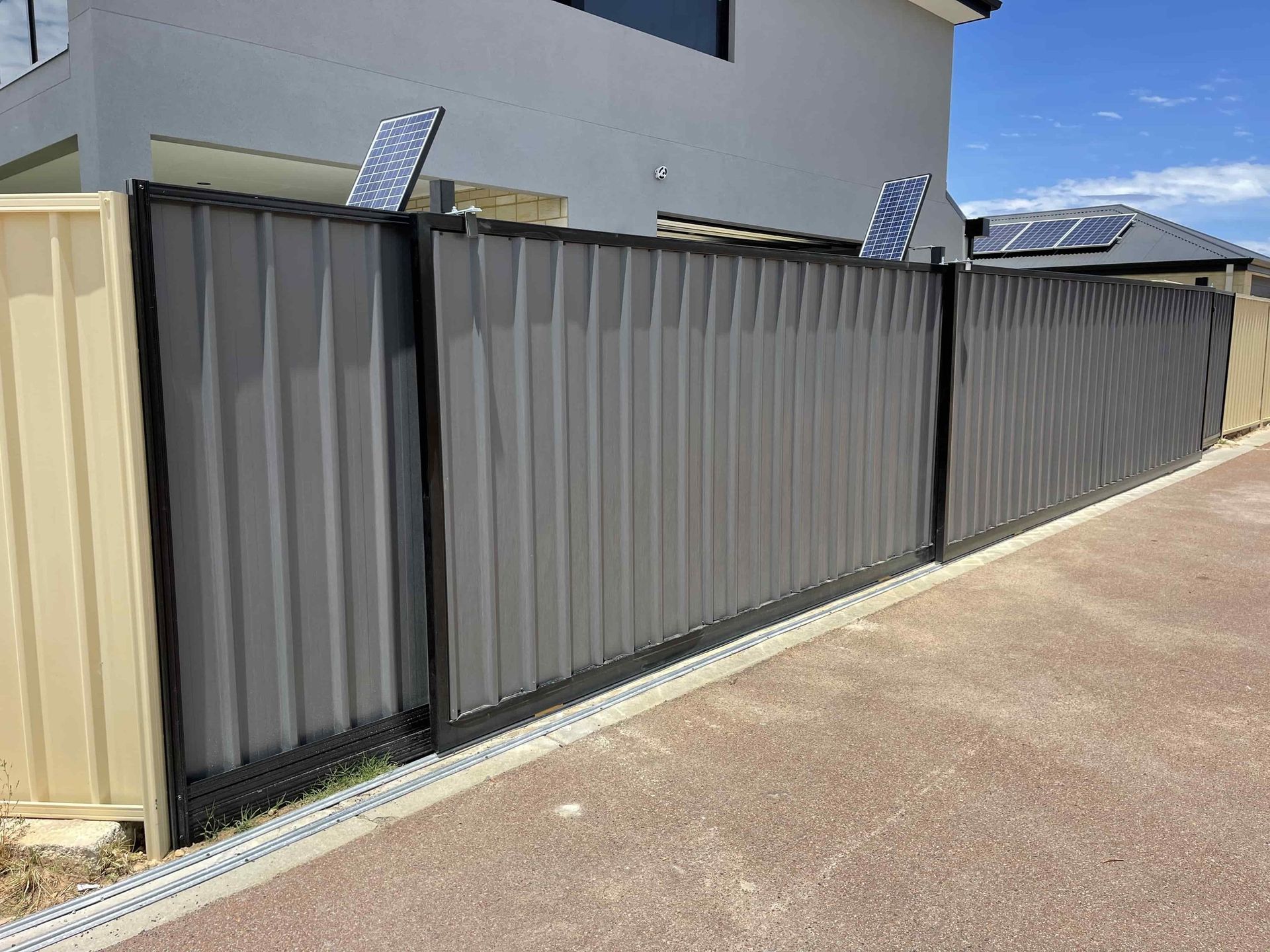 Long gray metal fence with slatted gates beside a driveway and beige wall in a residential area