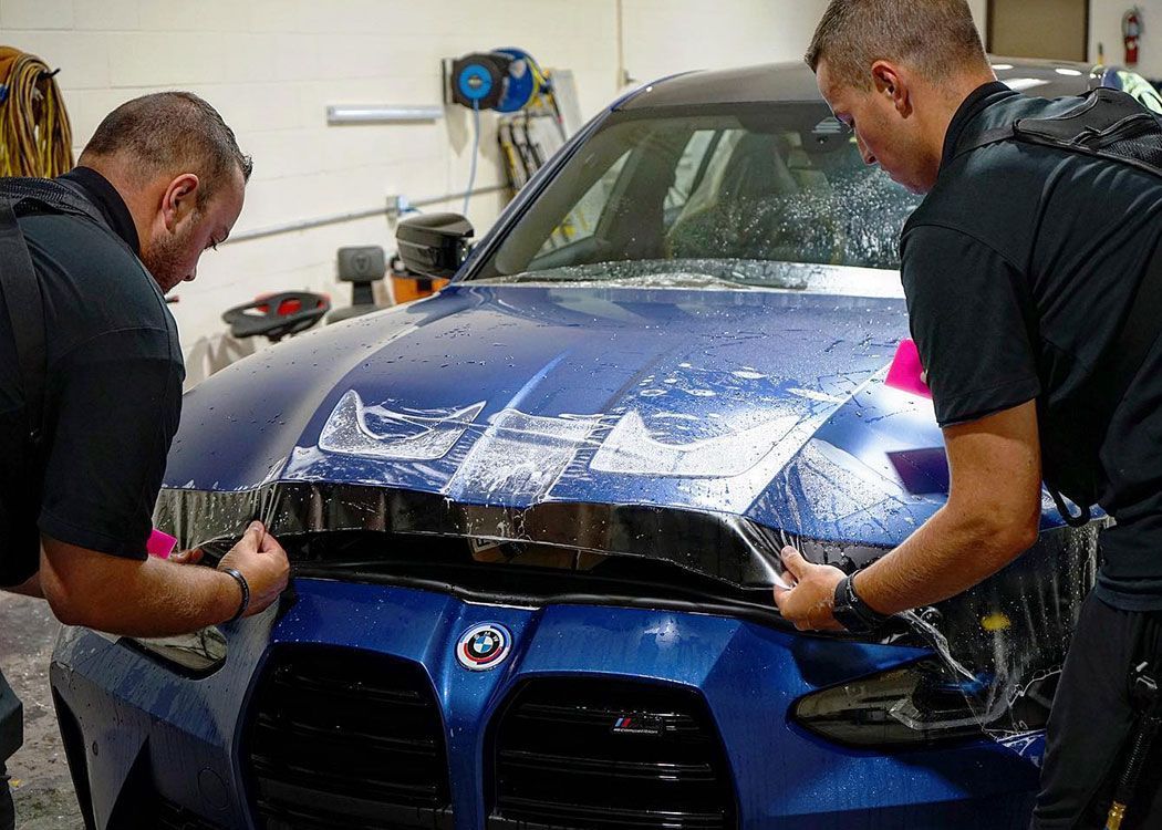 Two people applying protective film to the hood of a blue car in a garage.