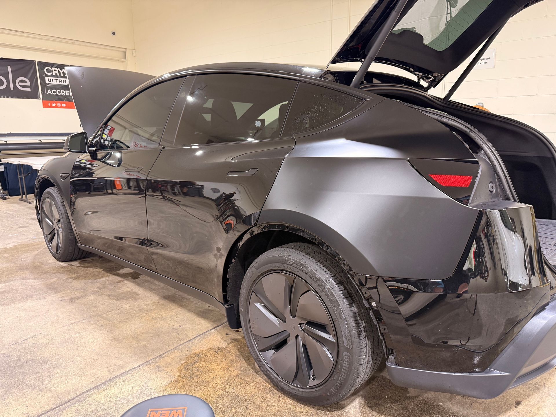 Black Tesla Model Y in a garage with rear hatch open and front wheel removed, showing damage on the side