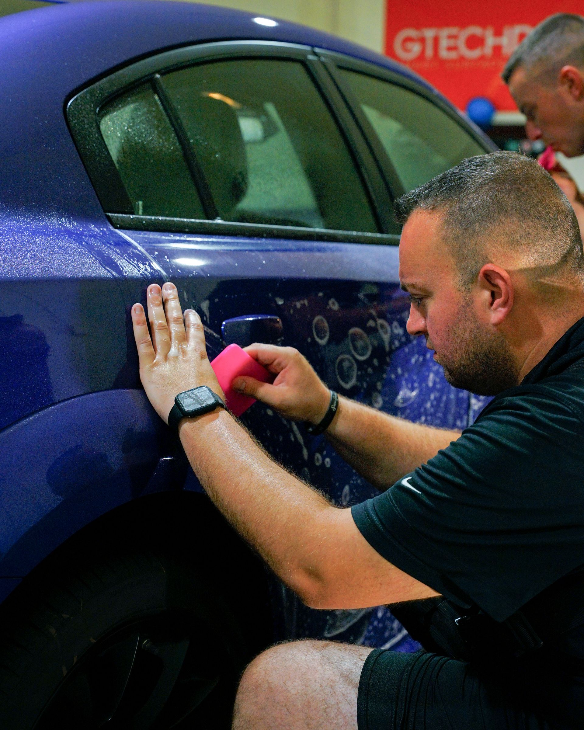 Man applying wrap to a blue car door with a pink squeegee. Indoors, focused expression.