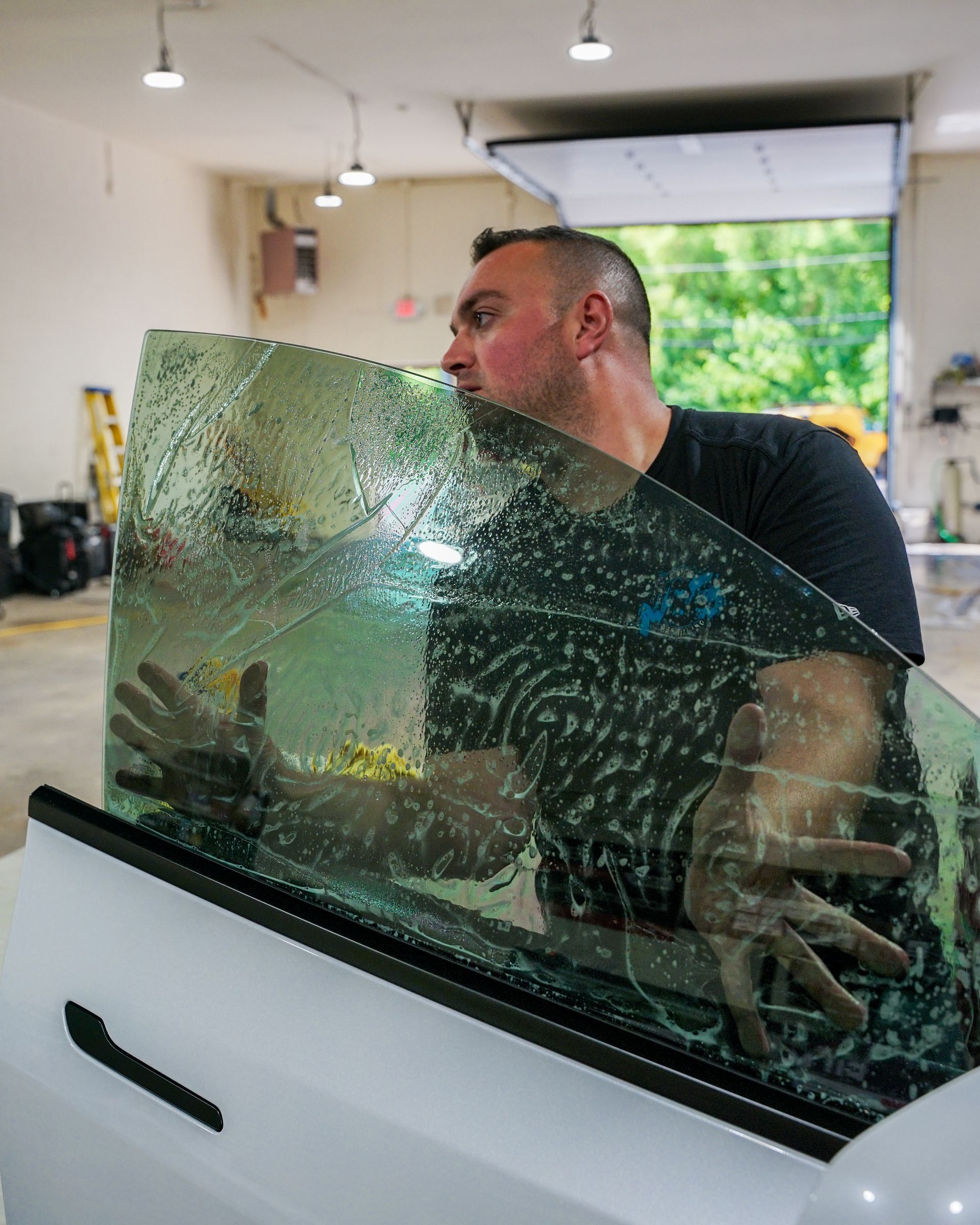 Man applying tinted film to a car window in a garage. He’s looking at the glass and using both hands.