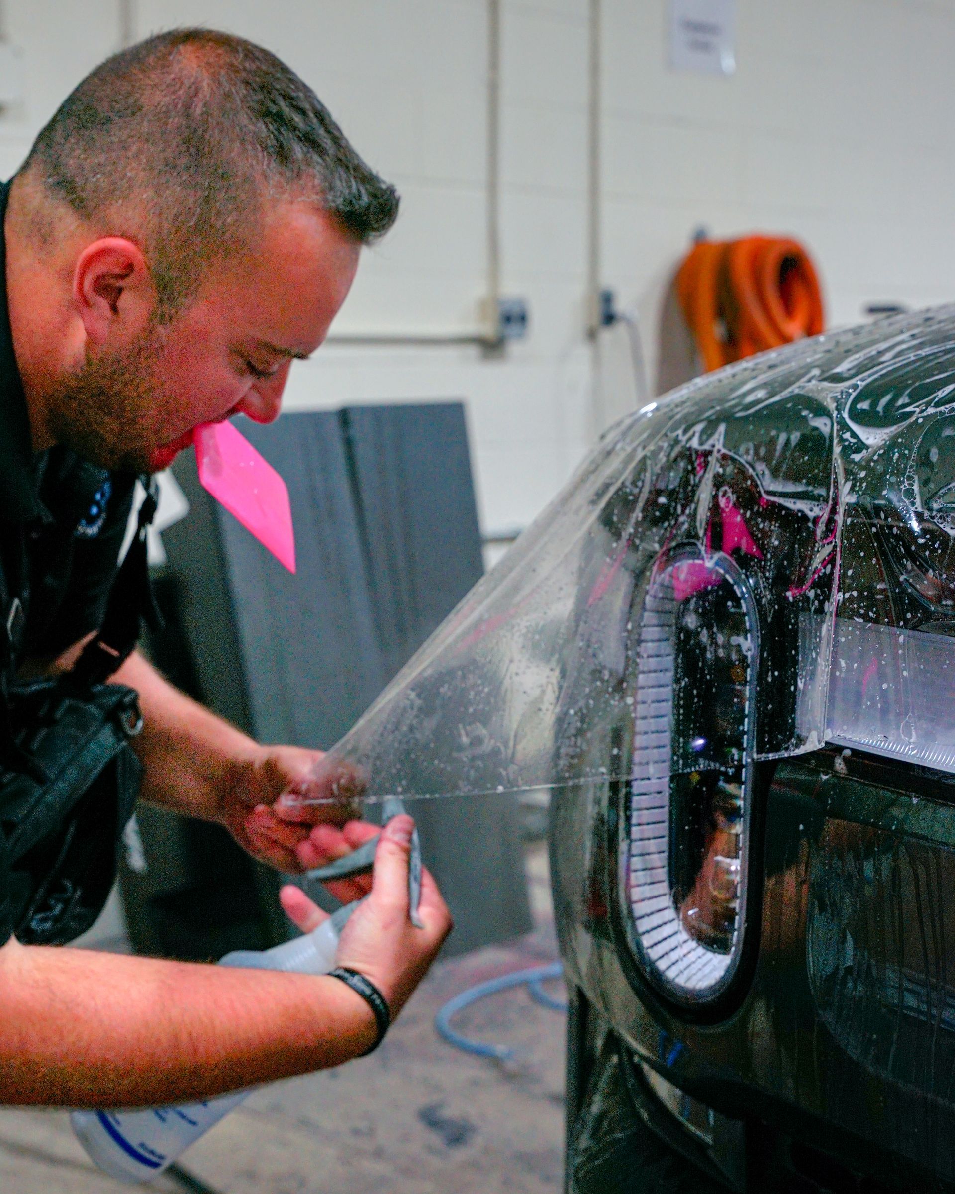 Man applying clear film to a car's taillight, using his hands and mouth, indoors.