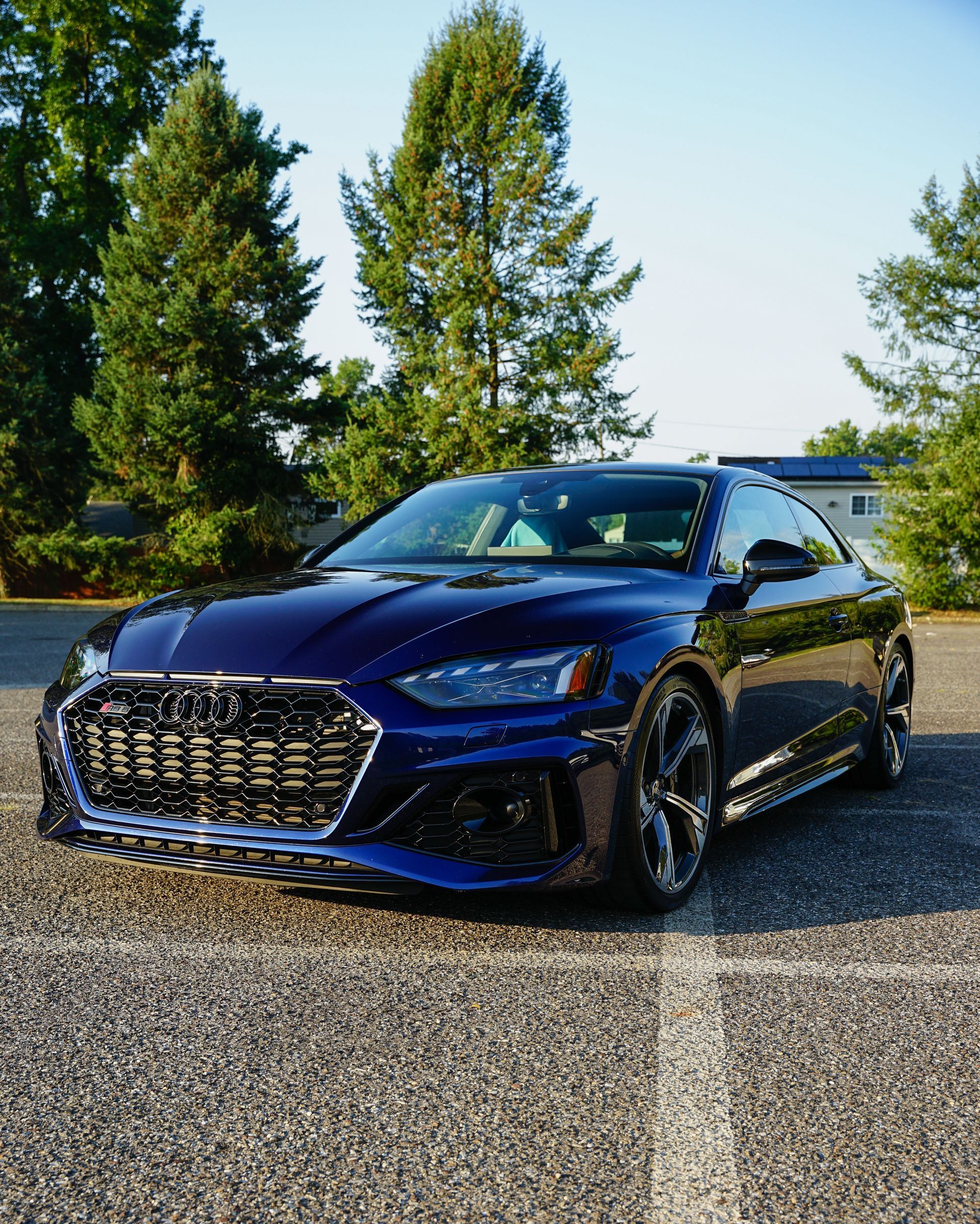 A dark blue Audi sports coupe parked on an asphalt lot with trees in the background under a clear sky.