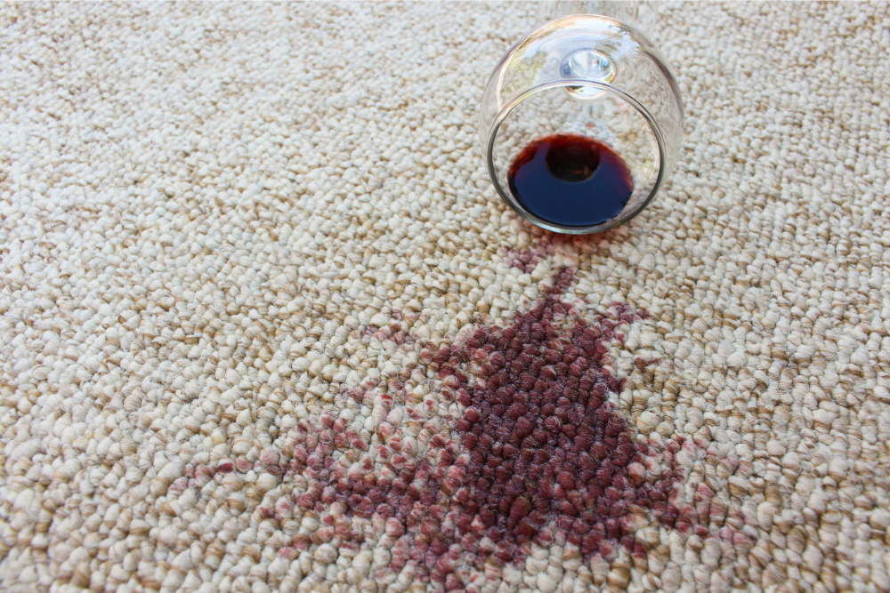 A glass of red wine spills onto a beige carpet, creating a dark red stain.