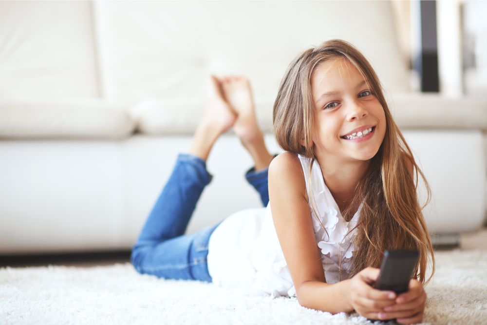 Girl lying on carpet with remote, smiling.