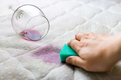 A hand sponges a red wine stain on a white mattress near a tipped-over wine glass. A hand sponges a red wine stain on a white mattress near a tipped-over wine glass.