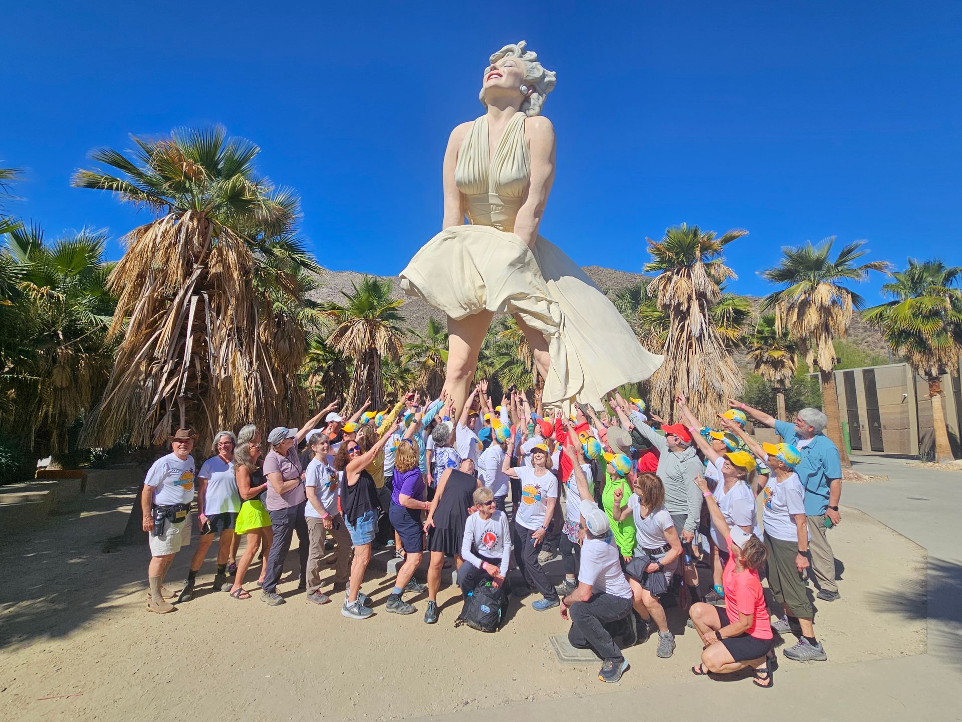People gather at Marilyn Monroe statue, pointing up. Palm trees and desert setting. Bright blue sky.