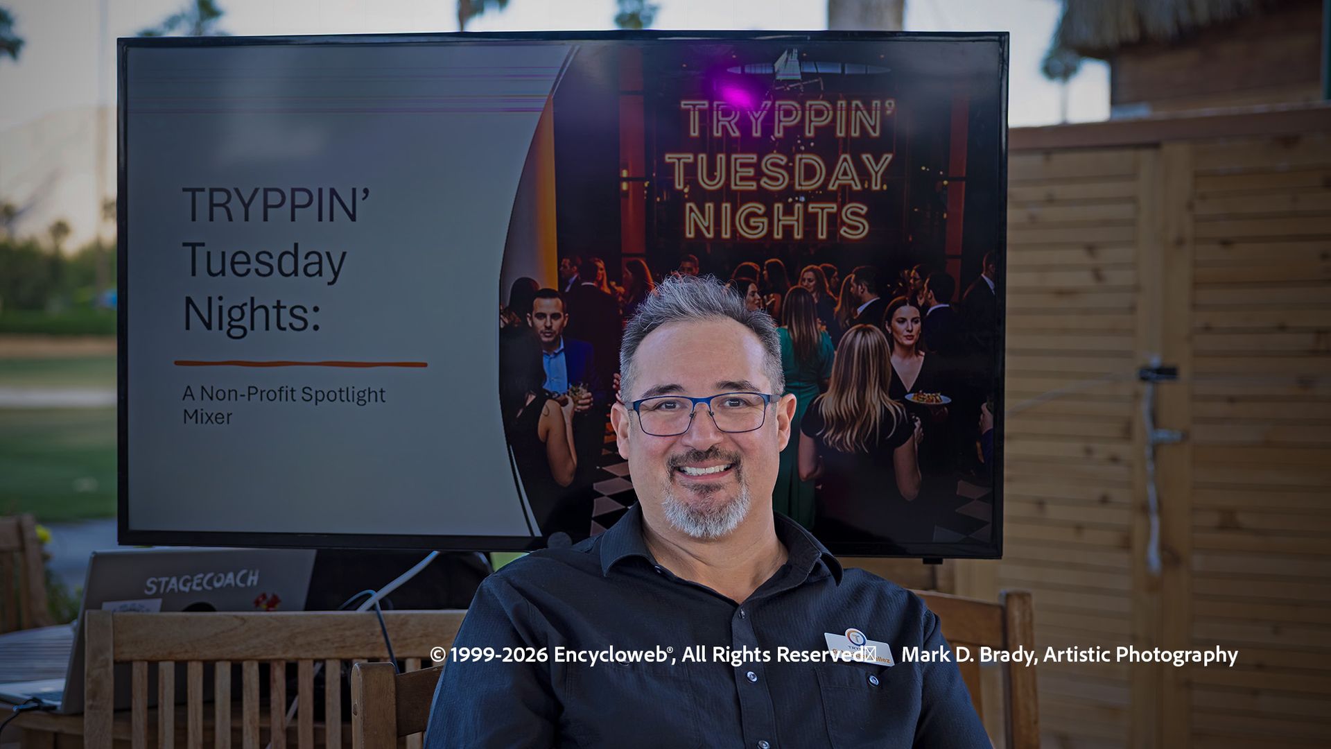 Man standing before a truck with a “Trippin’ Tuesday Nights” sign and neon-lit backdrop