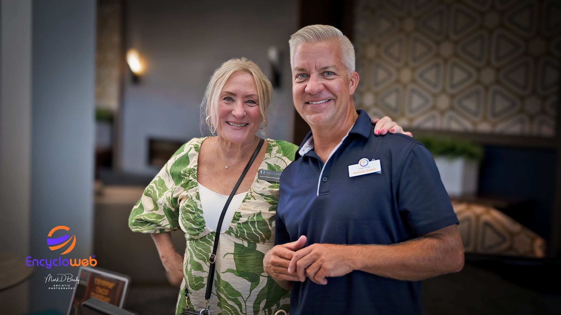 Two smiling people posing indoors, woman in green floral top and man in navy polo holding a drink.