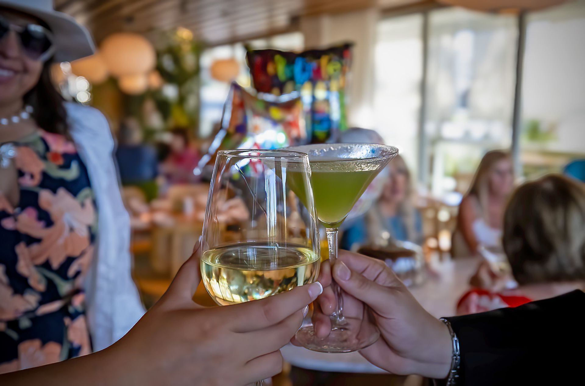 Hands clinking wine and a green cocktail, toasting in a restaurant with others.