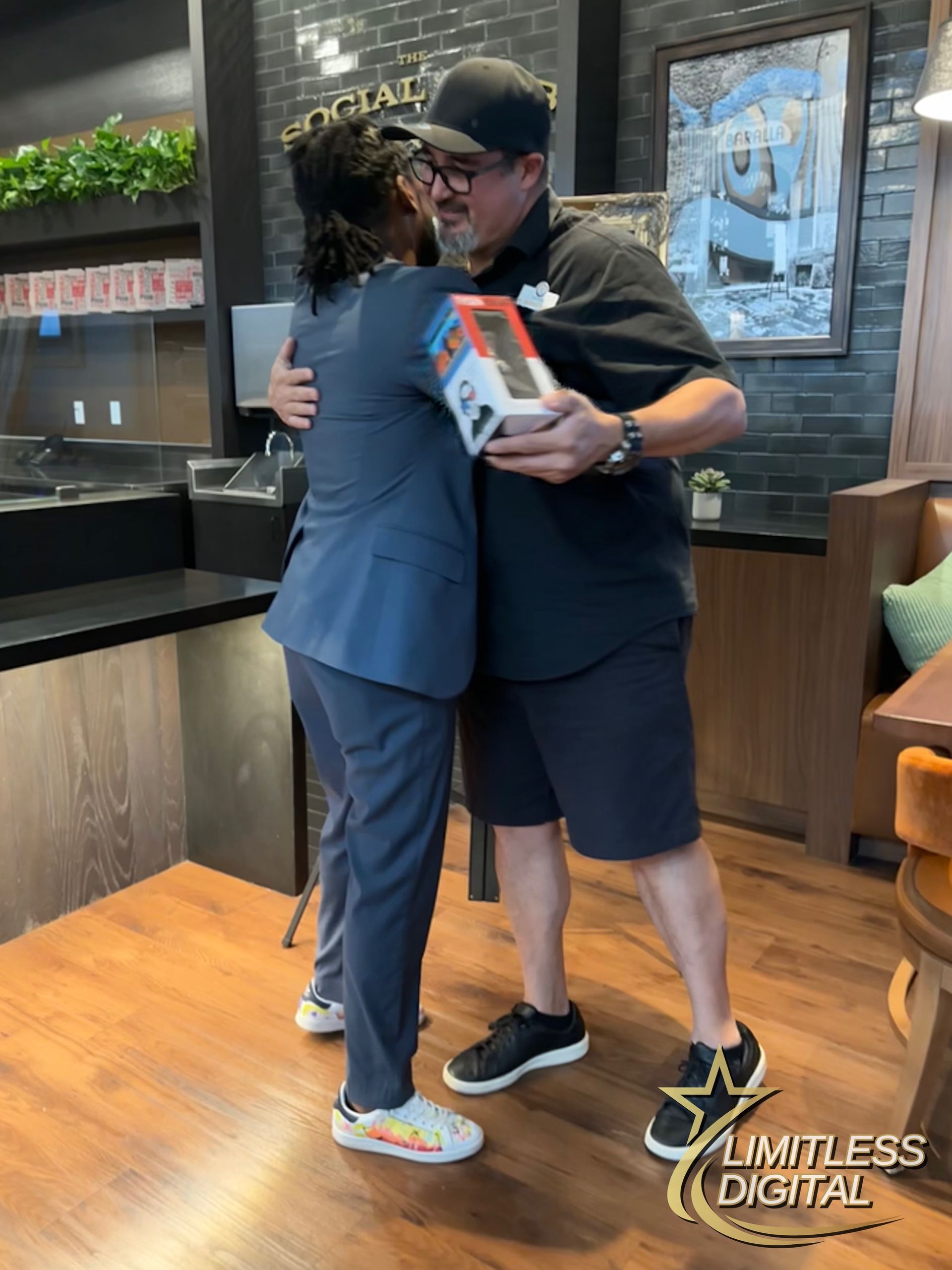 Two people hugging indoors, one holding a boxed item, with wood flooring and a dark wall behind them.
