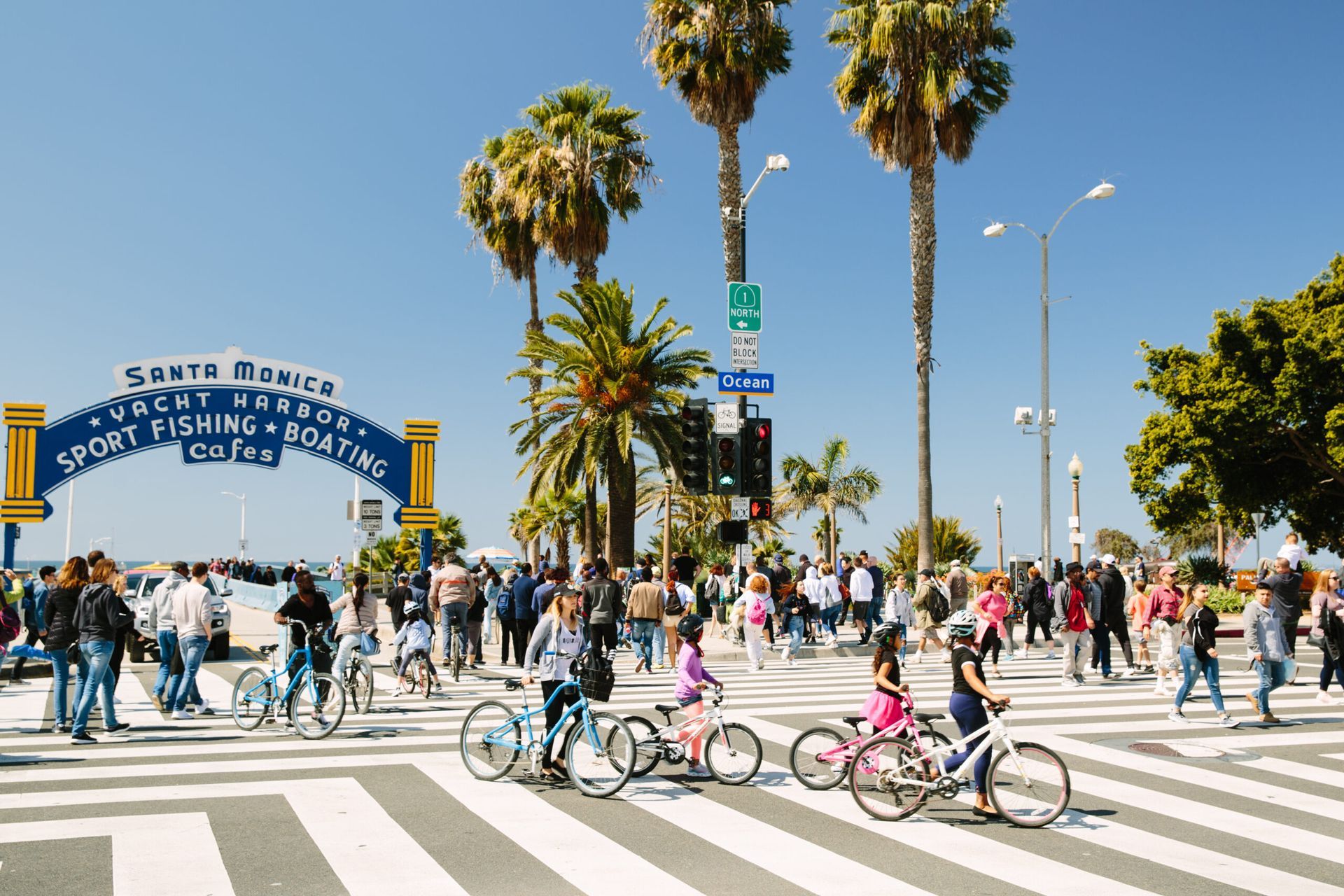 People crosswalk at Santa Monica pier with bikes, blue sky, palm trees, and the pier entrance arch.