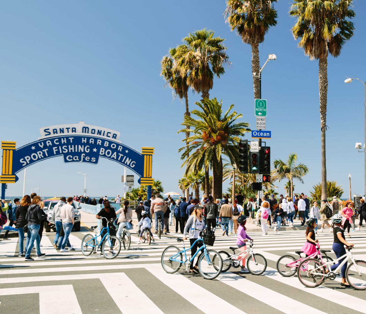 Santa Monica pier entrance with cyclists crossing a busy crosswalk. Palm trees and a blue sky are visible.
