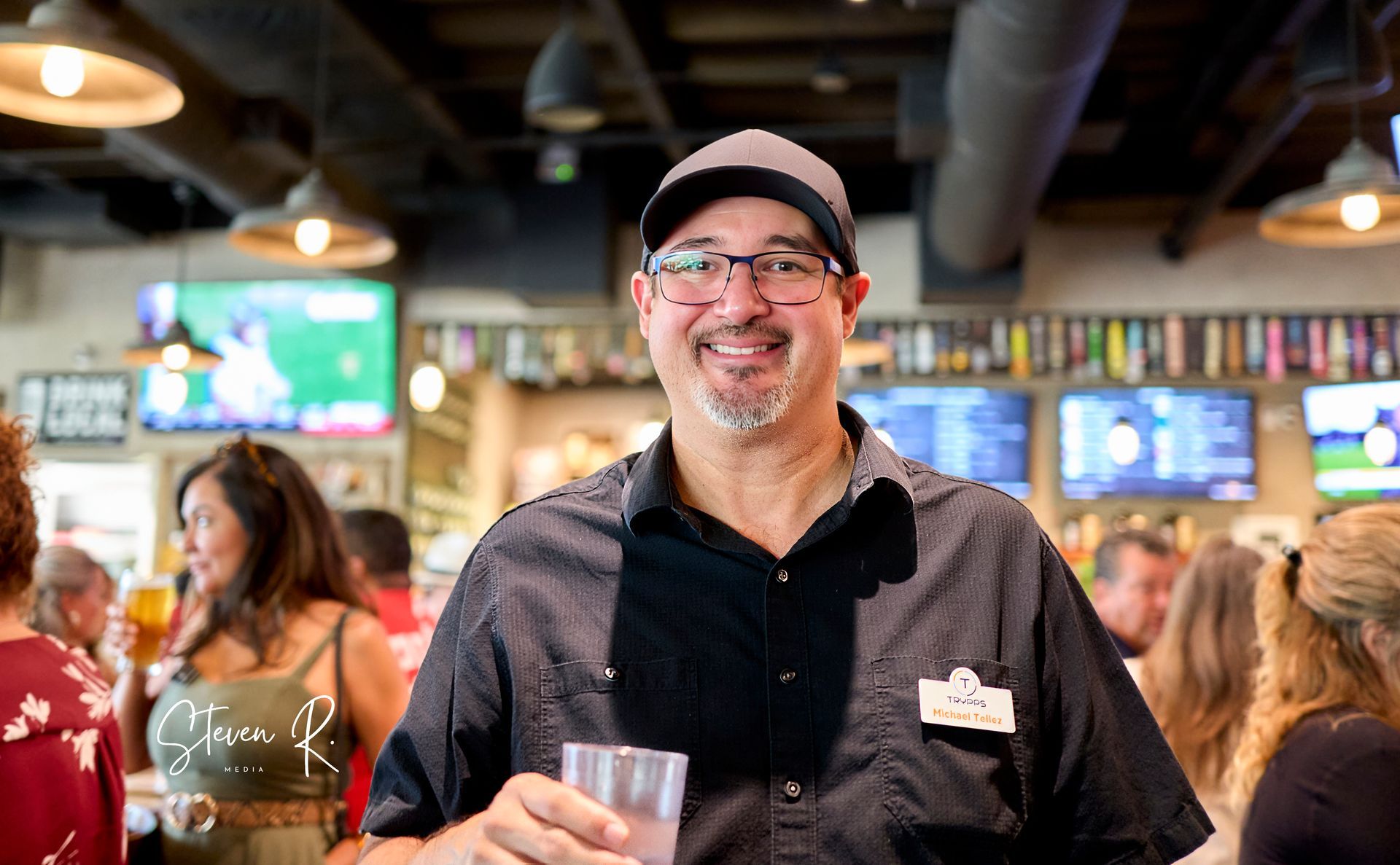 Man smiling in a bar, holding a drink, wearing a hat and glasses, surrounded by people and TVs.