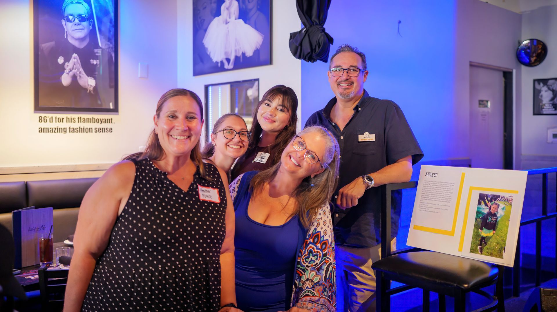 Group of five people smiling inside a bar with blue lighting and a picture of Elton John on the wall.