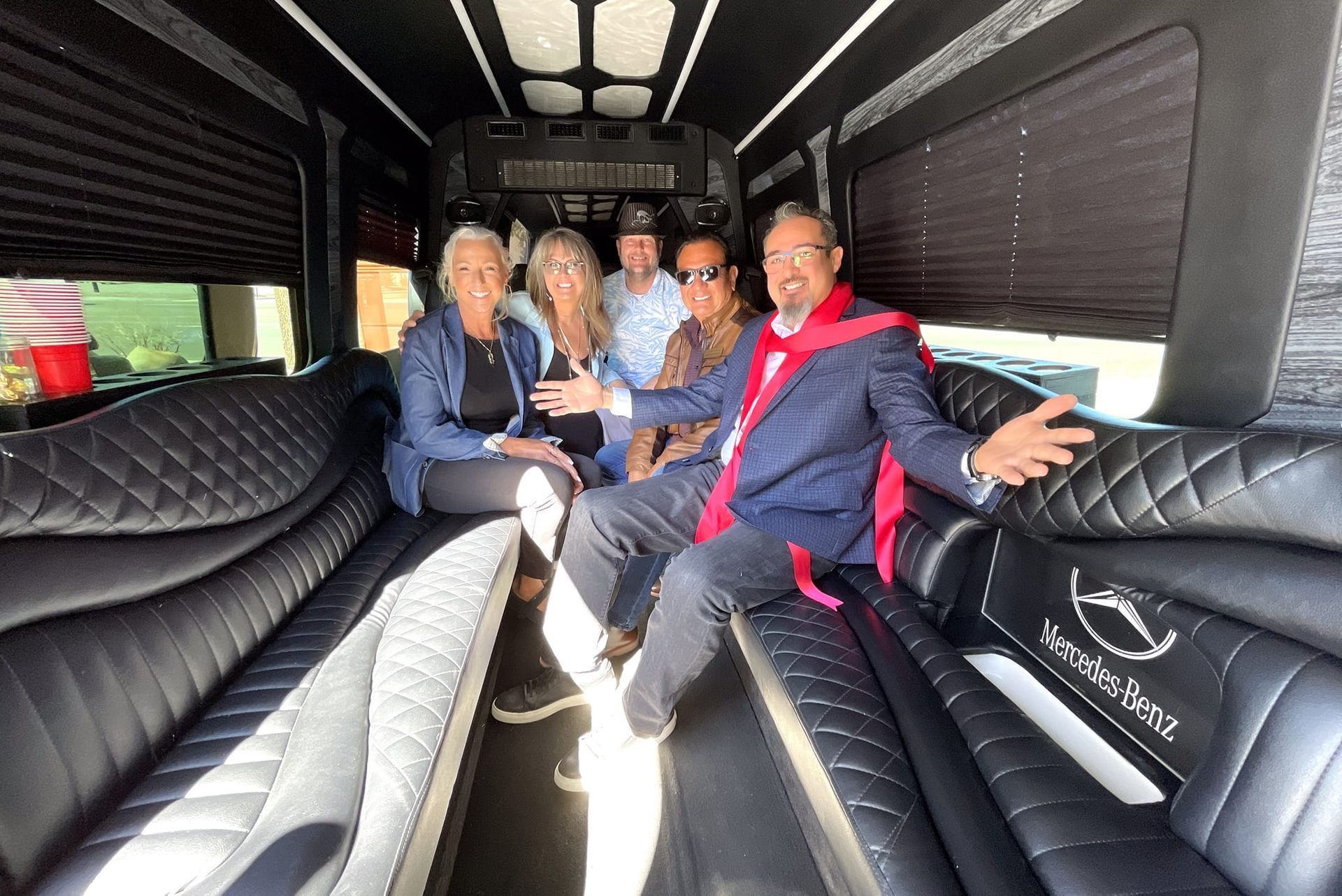 People inside a luxury van with black quilted seats. Man in blue blazer, red scarf smiles, arms open. Others smile.