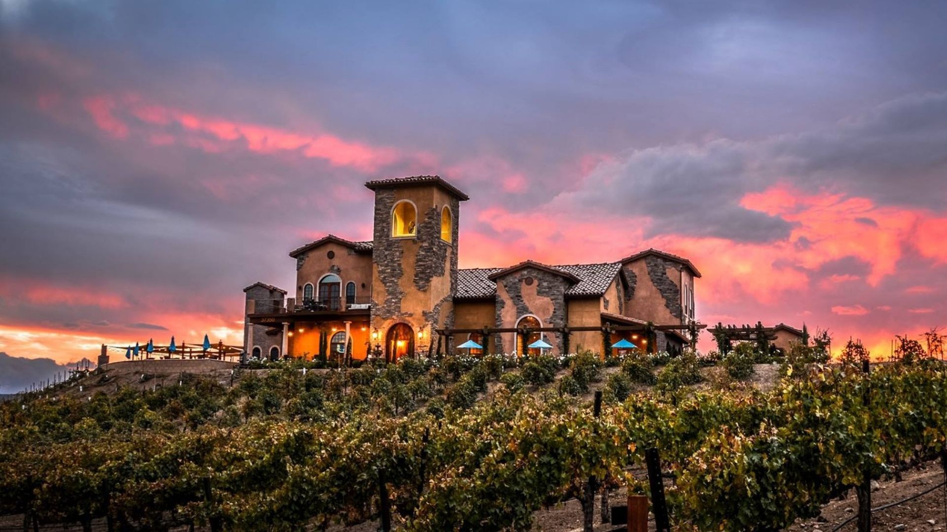 Winery at sunset, building with tower sits atop a hill, surrounded by vineyards, against a colorful sky.