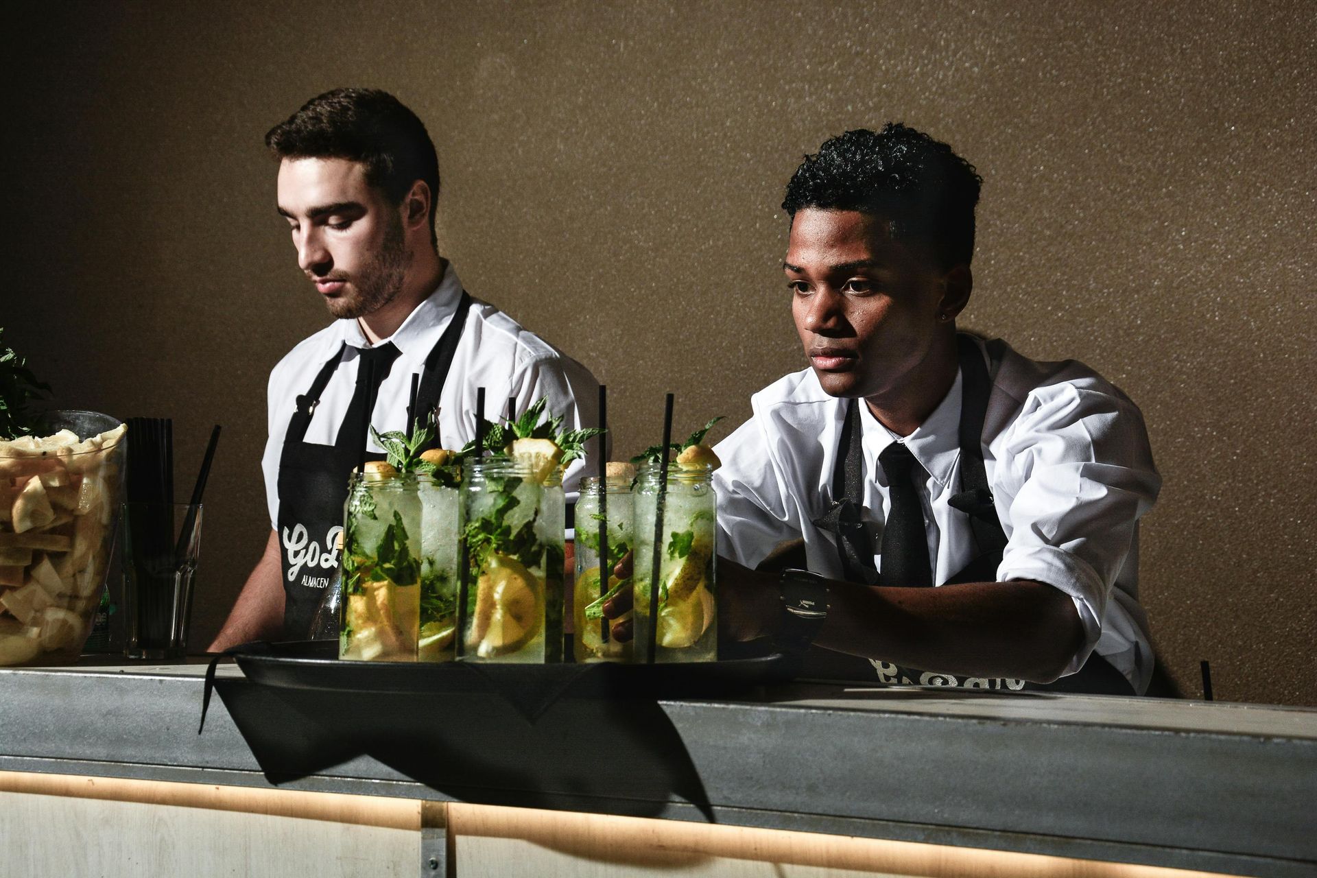 Two bartenders preparing cocktails behind a bar, drinks in the foreground.