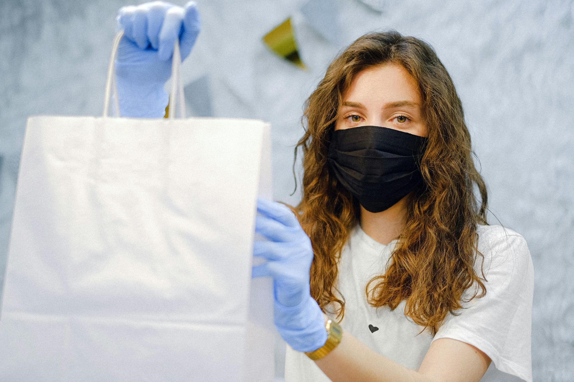 Woman in mask and gloves holding white paper bag.