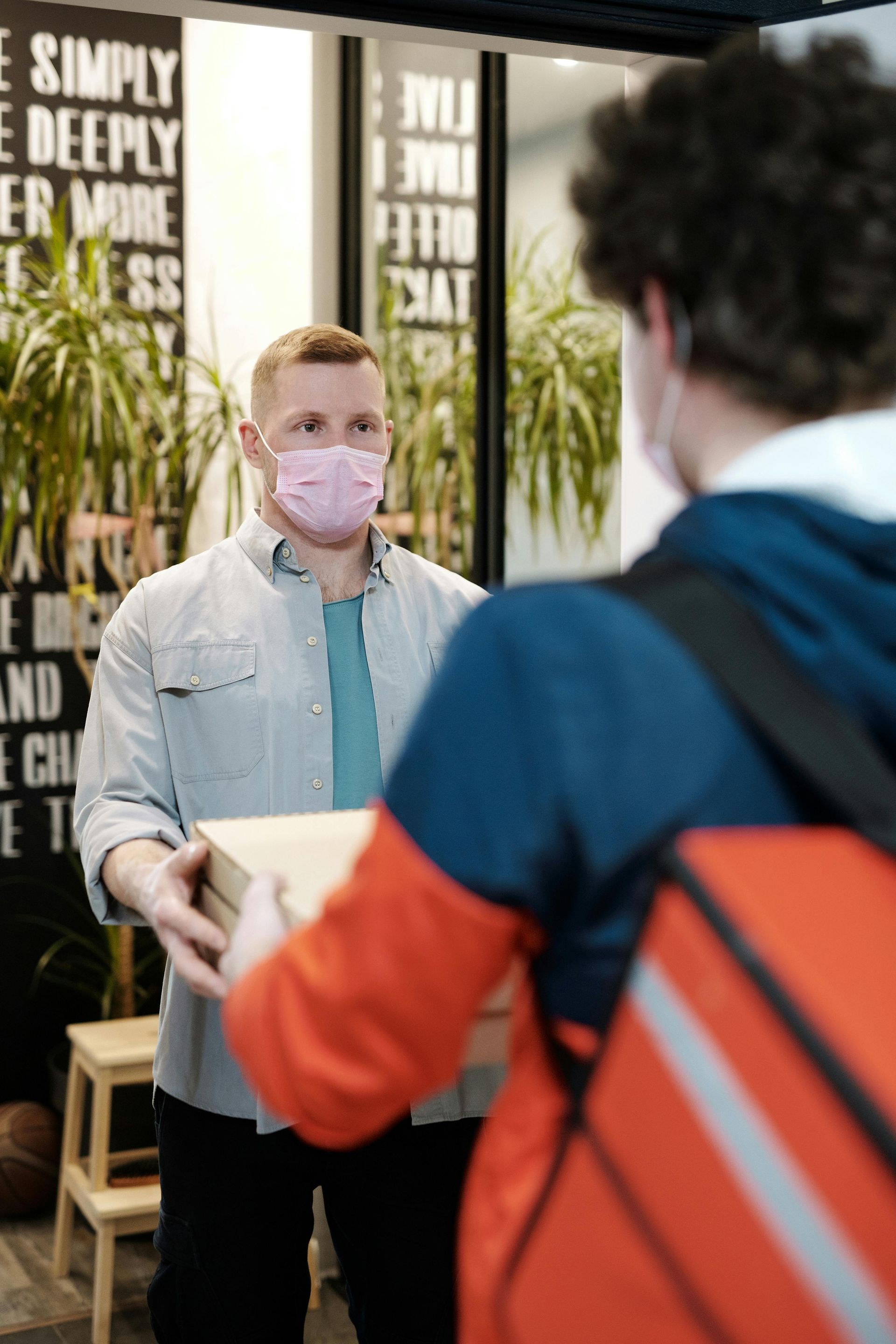 Man in pink mask receiving a delivery from a person with a red delivery bag.