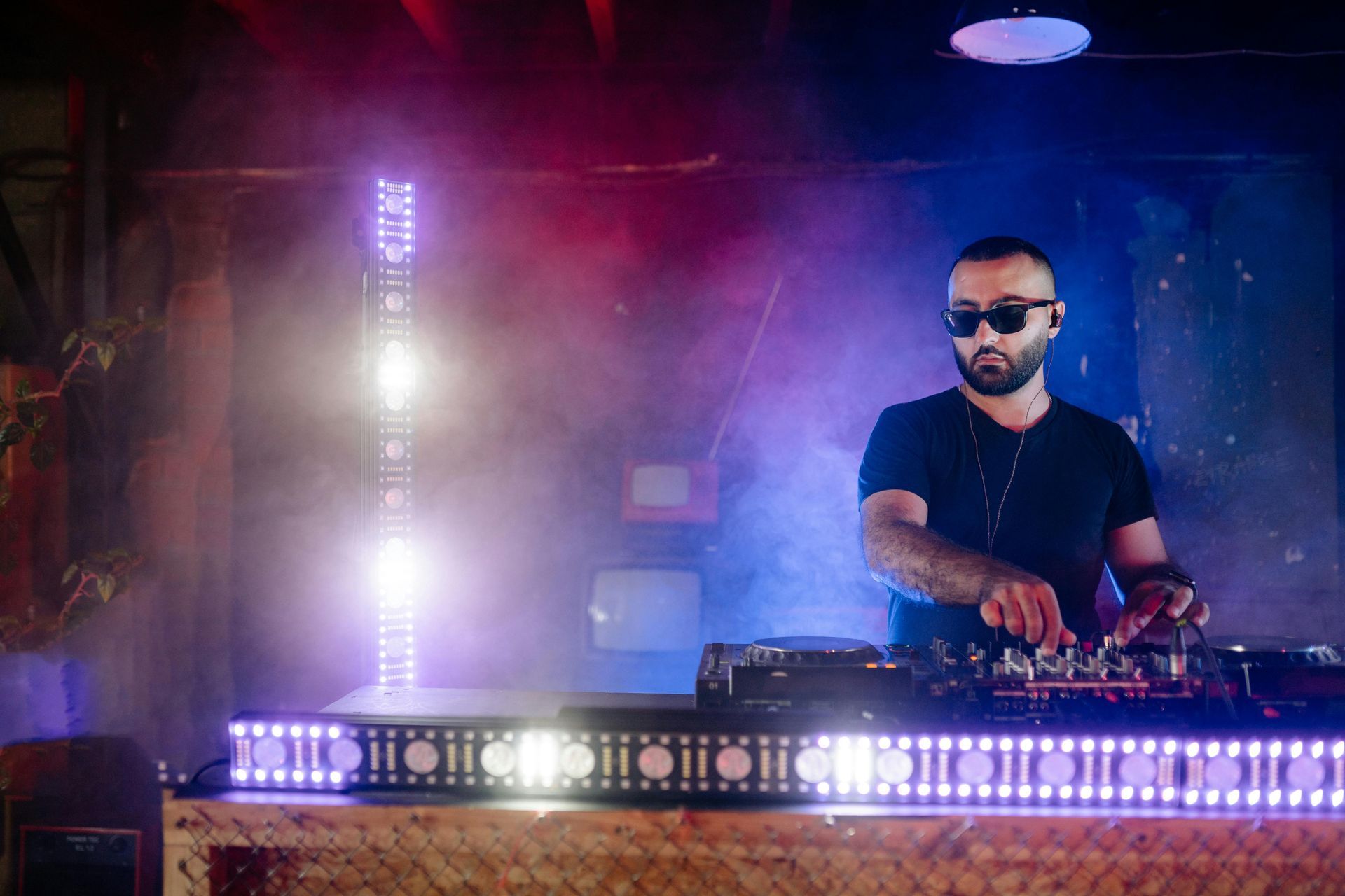 DJ at a club, wearing sunglasses, mixing on equipment. Stage lit with purple, blue, and white lights.