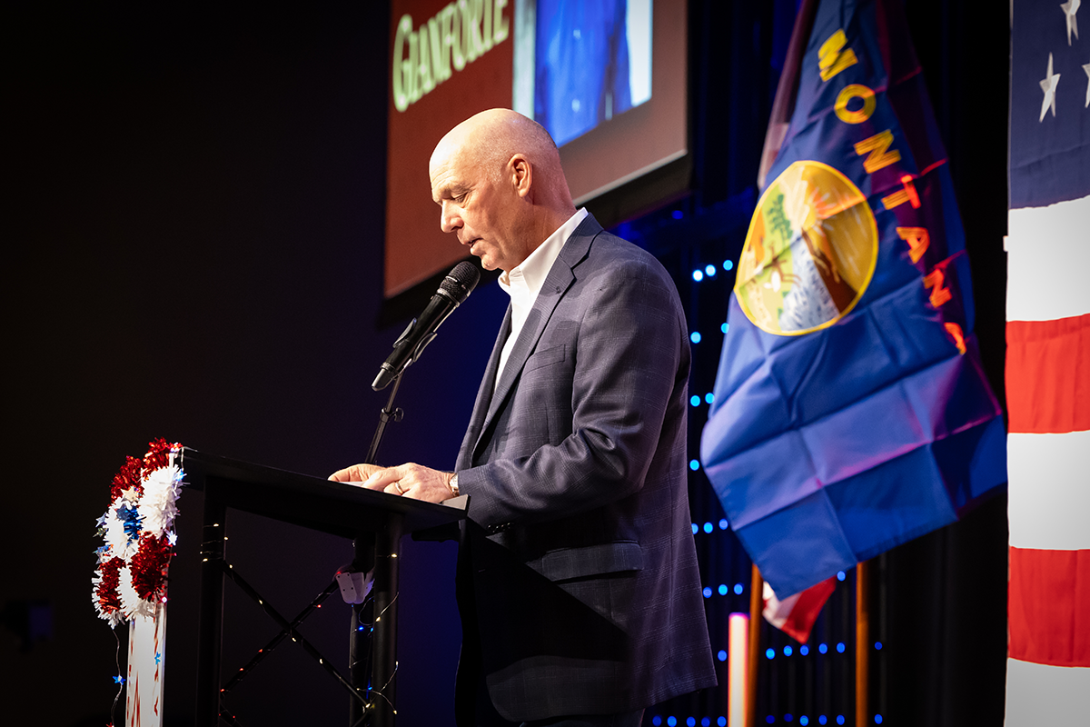 Speaker at podium on stage beside a blue flag, addressing an audience under stage lighting.