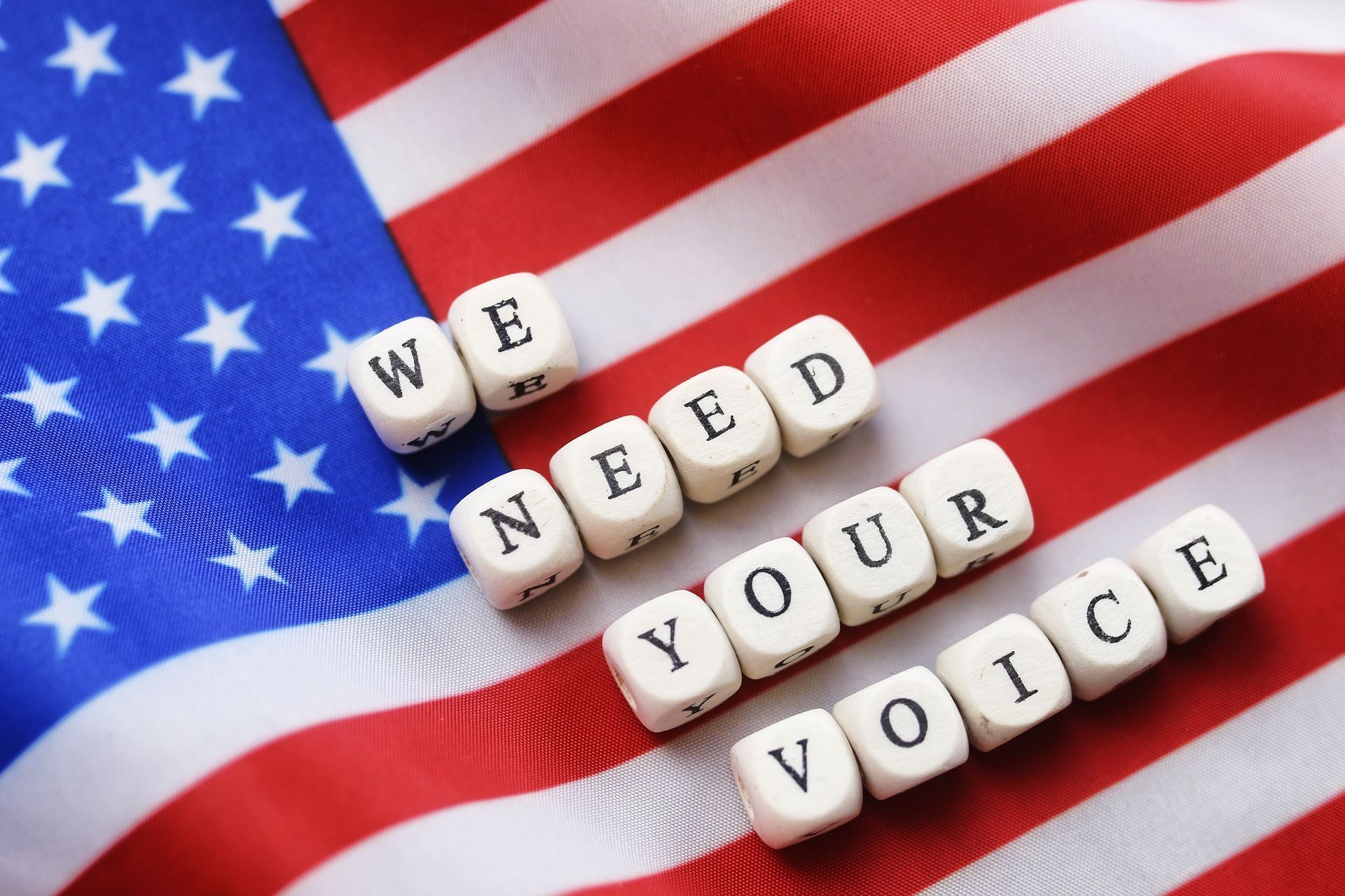 White letter tiles spelling “WE NEED YOUR VOICE” on a waving American flag background