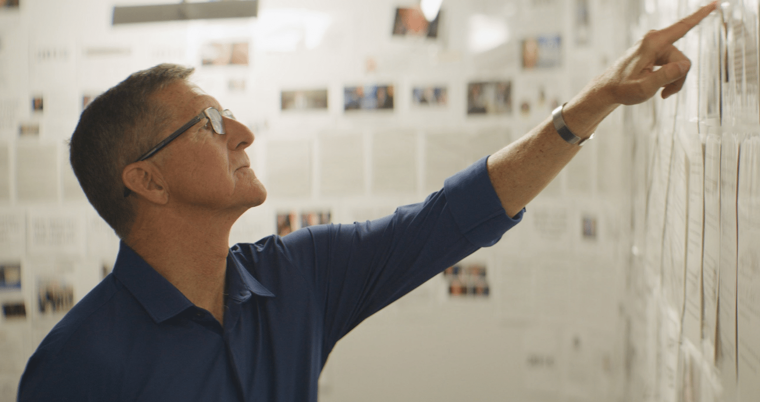Man in glasses pointing at a wall of pinned notes and photos in a bright office