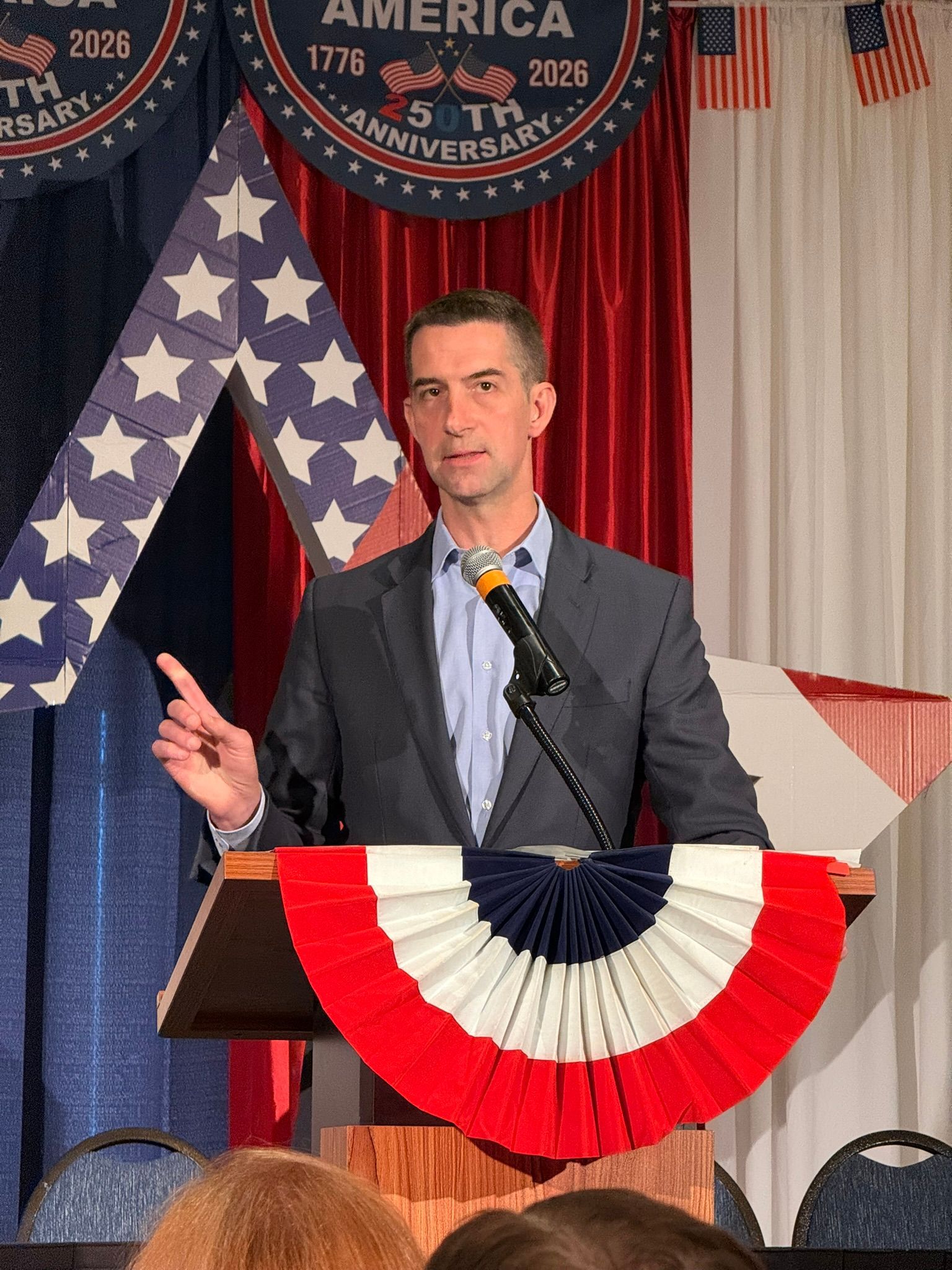 Man speaking at a podium with red, white, and blue bunting in front of patriotic flags and a curtain backdrop