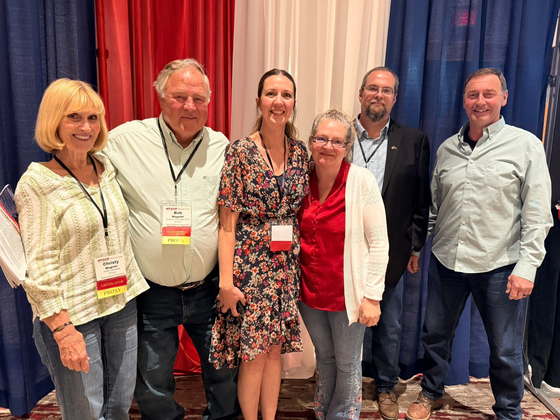 Six people standing together at a conference, smiling in front of red and beige curtains