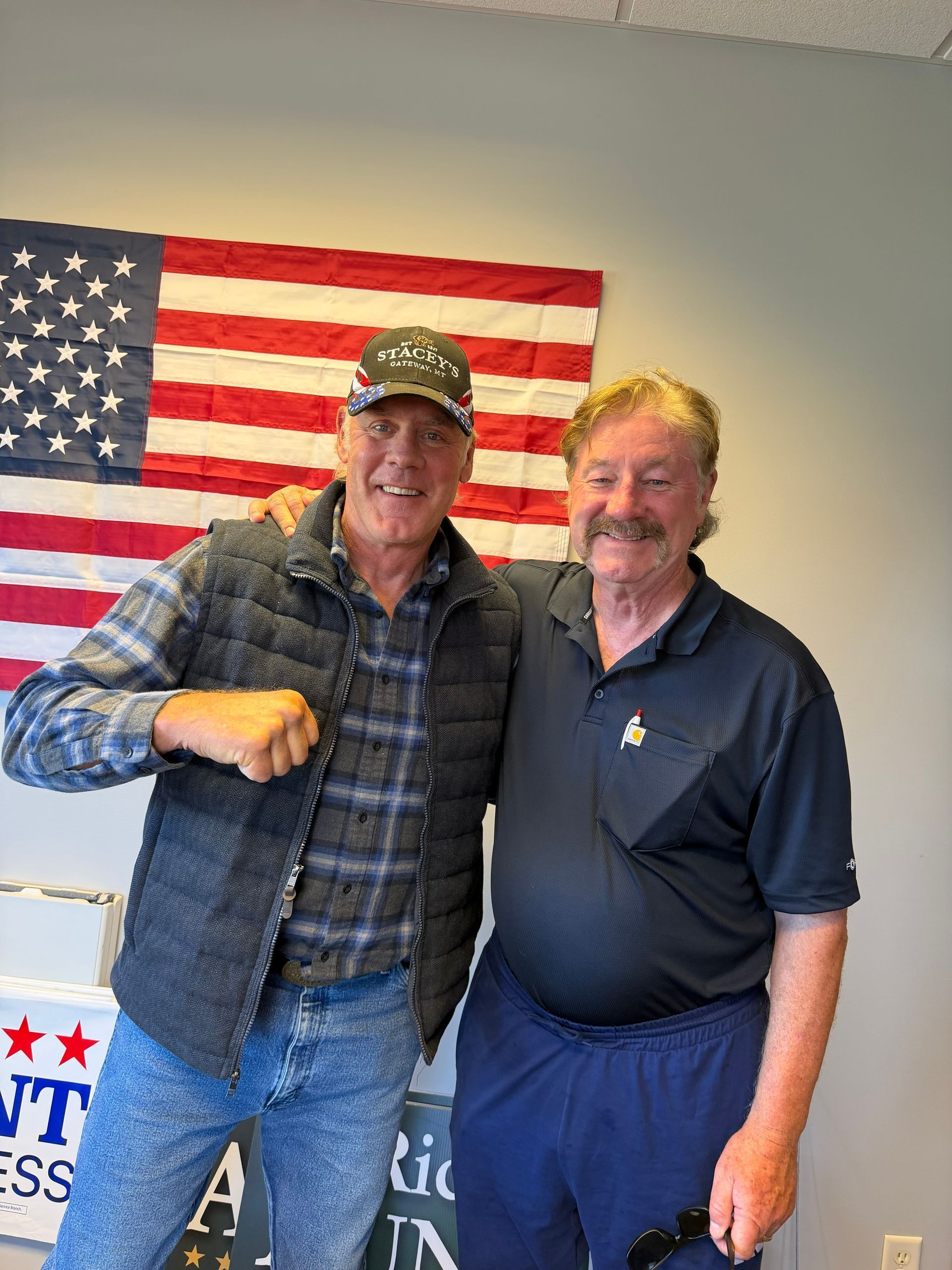 Two men smiling and posing indoors in front of a large American flag