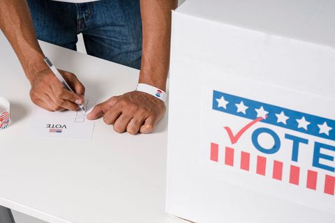 Person signing a ballot beside a white ballot box labeled “VOTE” with stars and stripes