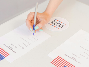 Hand writing on a worksheet next to a “VOTE” sticker and U.S. flag graphics