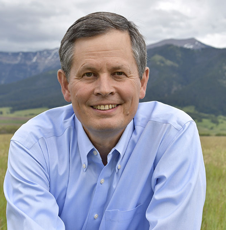 Man in a light blue shirt smiles outdoors with mountains in the background