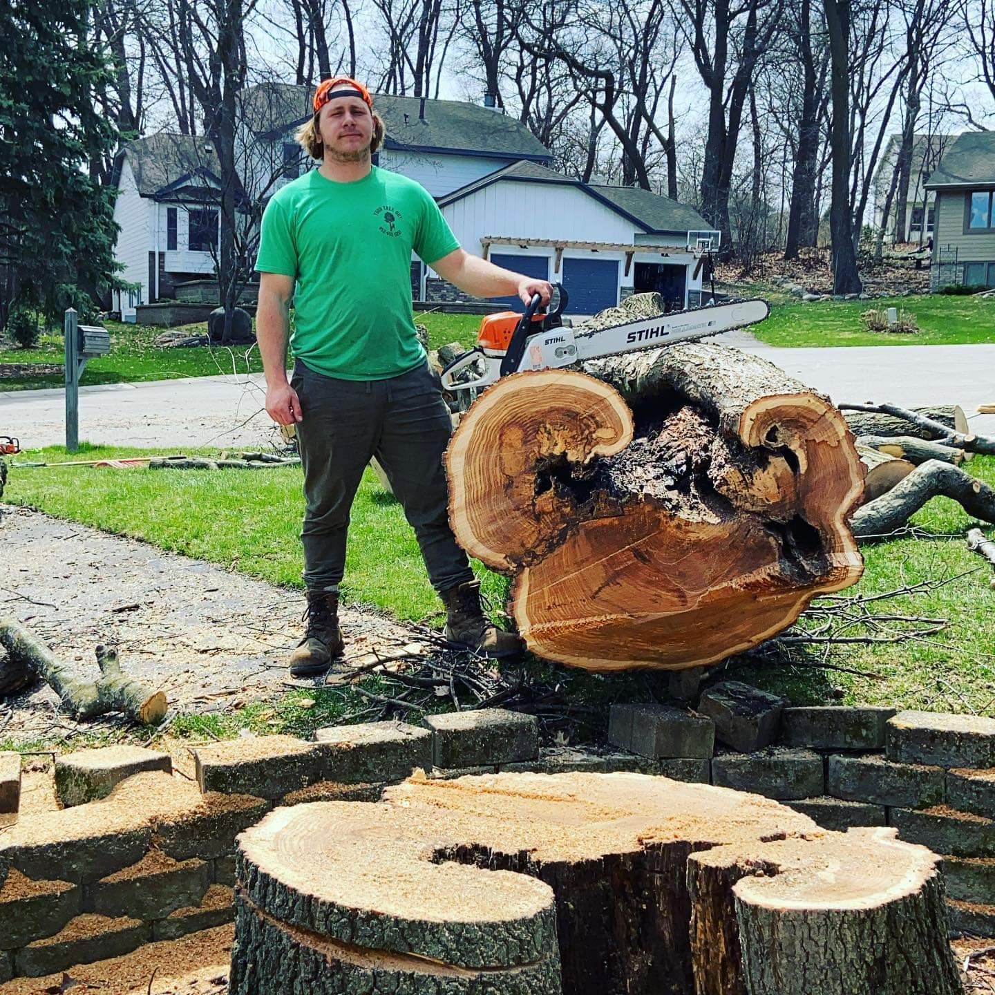 A man is standing next to a large tree stump with a chainsaw - Shakope, MN - Your Tree Guy, LLC