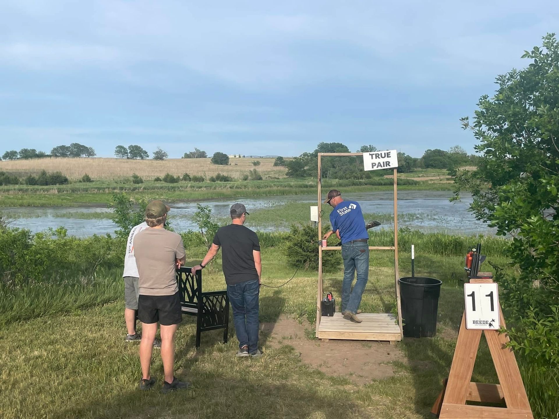 Man aiming a shotgun at a clay target, standing in a field with hay bales.