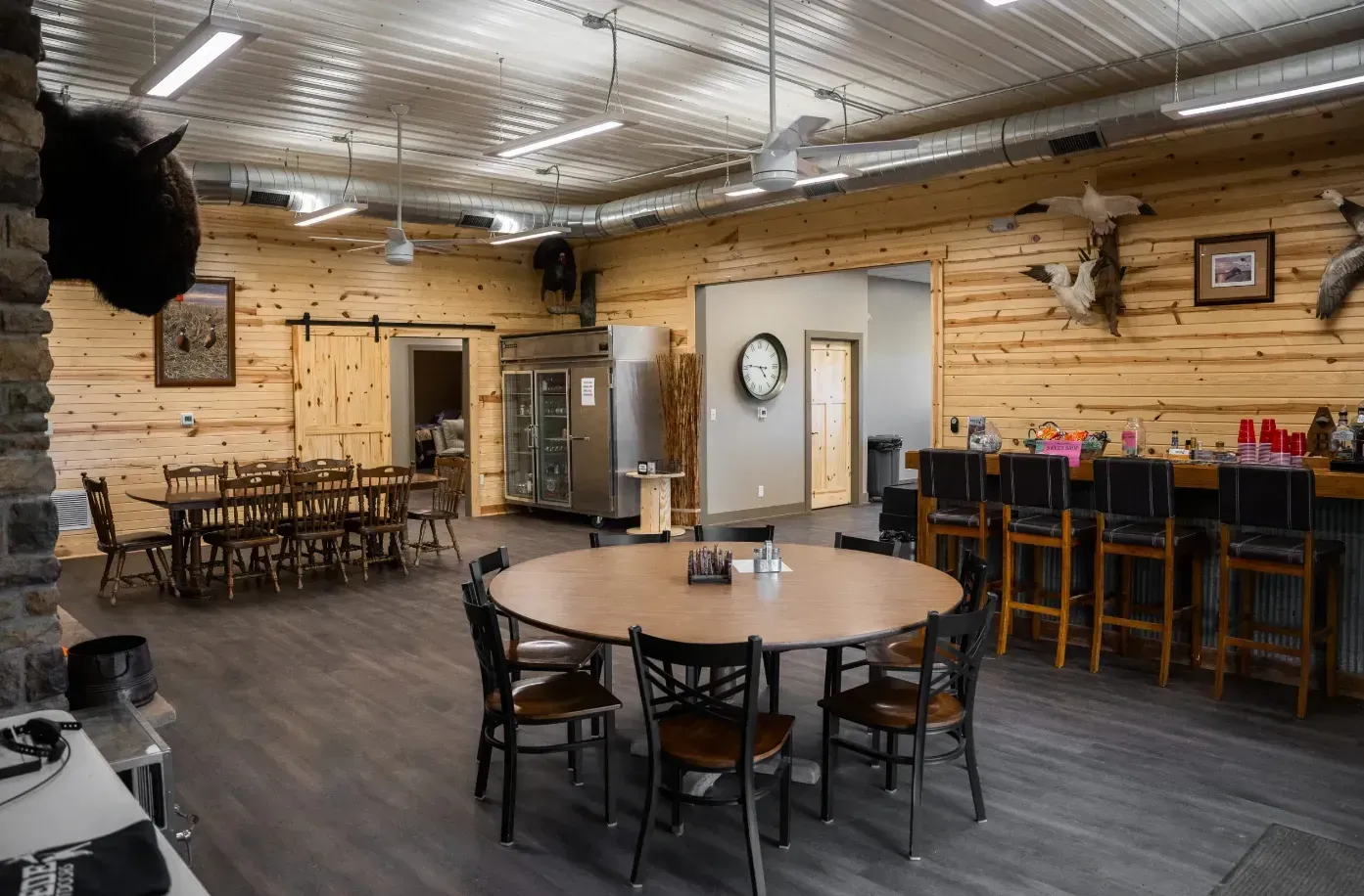 Rustic interior: bar, round table with chairs, dining tables, taxidermied animal heads, wooden walls, concrete floor.