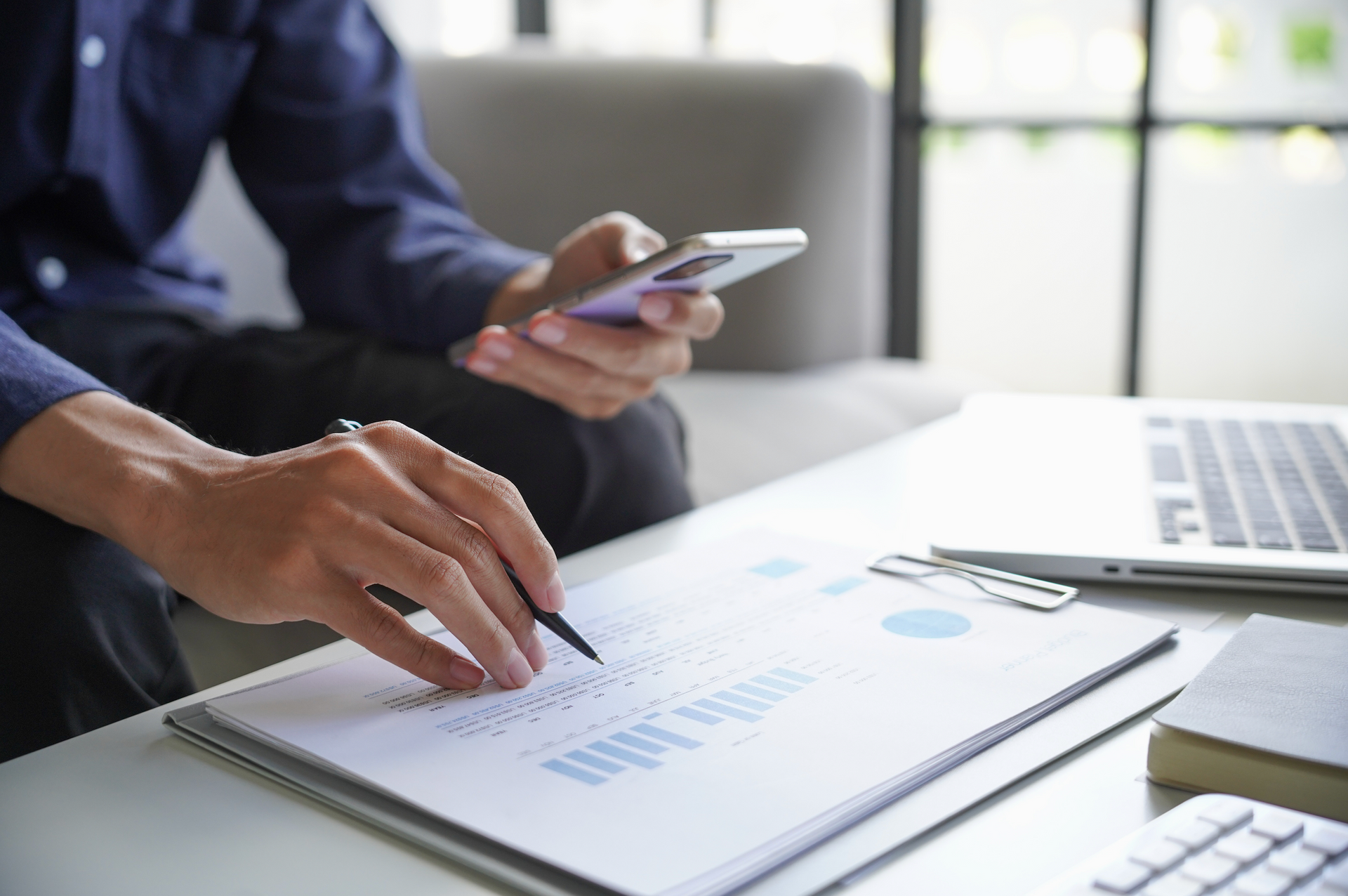 Person reviewing financial documents, holding a phone and pen, near a laptop and window.