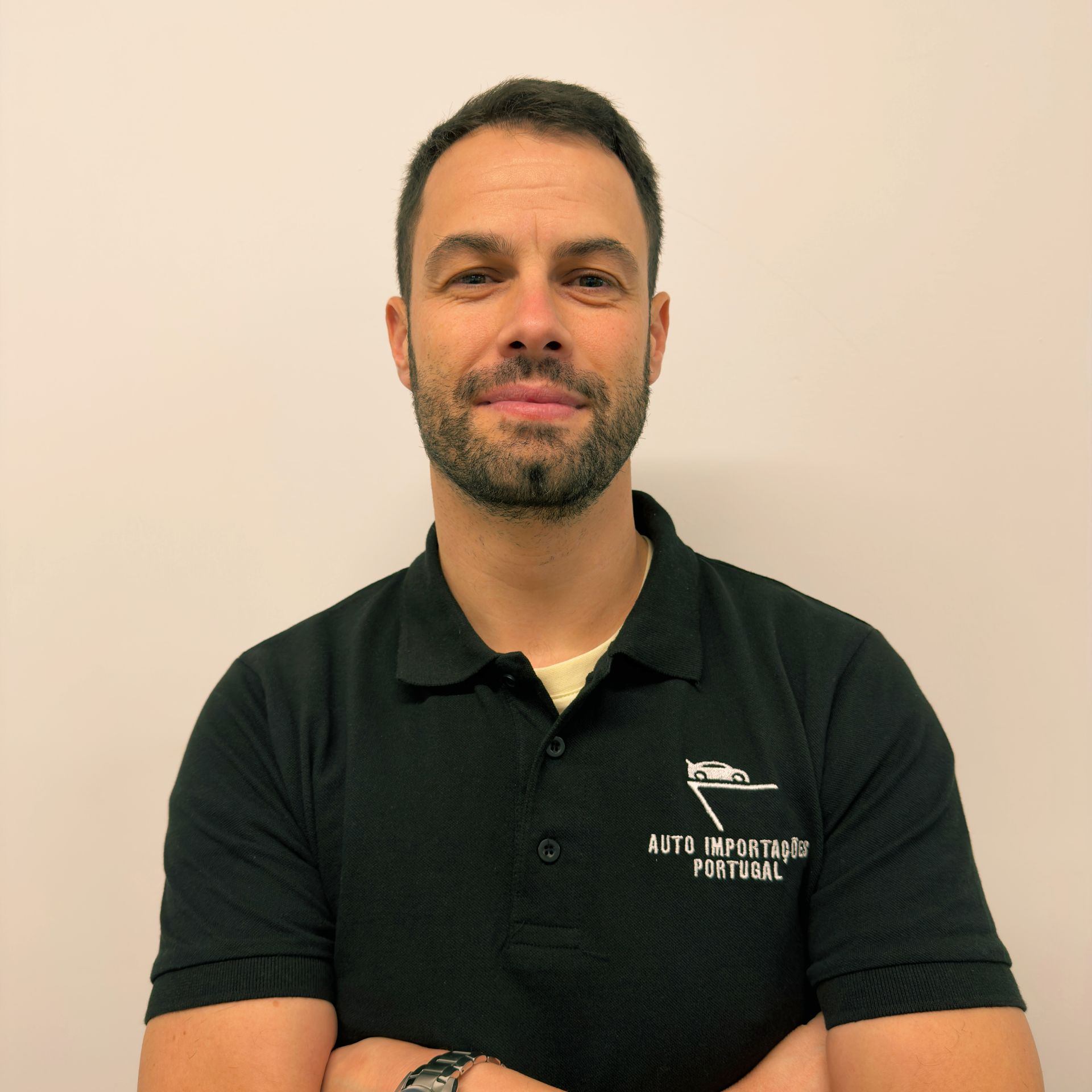 Homem vestindo camisa polo preta, em pé contra uma parede bege, de frente para a câmera com os braços cruzados.