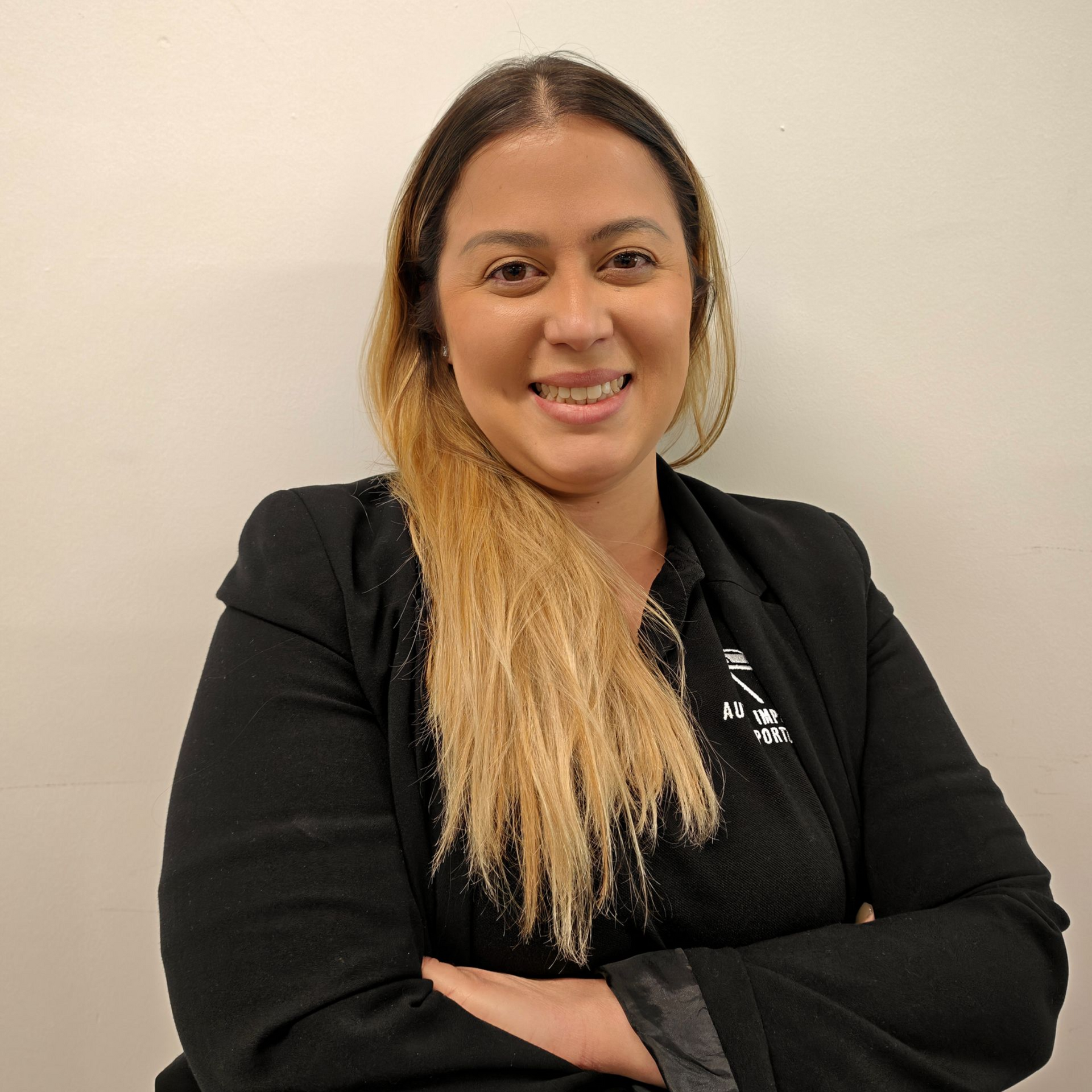 Mulher sorridente de blazer preto com os braços cruzados encostada em uma parede bege lisa.