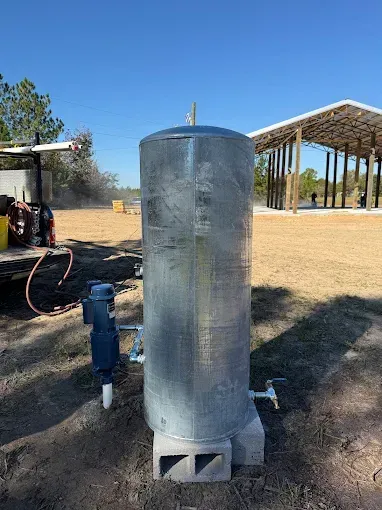 A vertical, galvanized steel water pressure tank sits on concrete blocks outdoors near a partially built wooden structure.