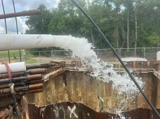 Water forcefully discharging from a white PVC pipe into a metal-walled construction excavation pit.