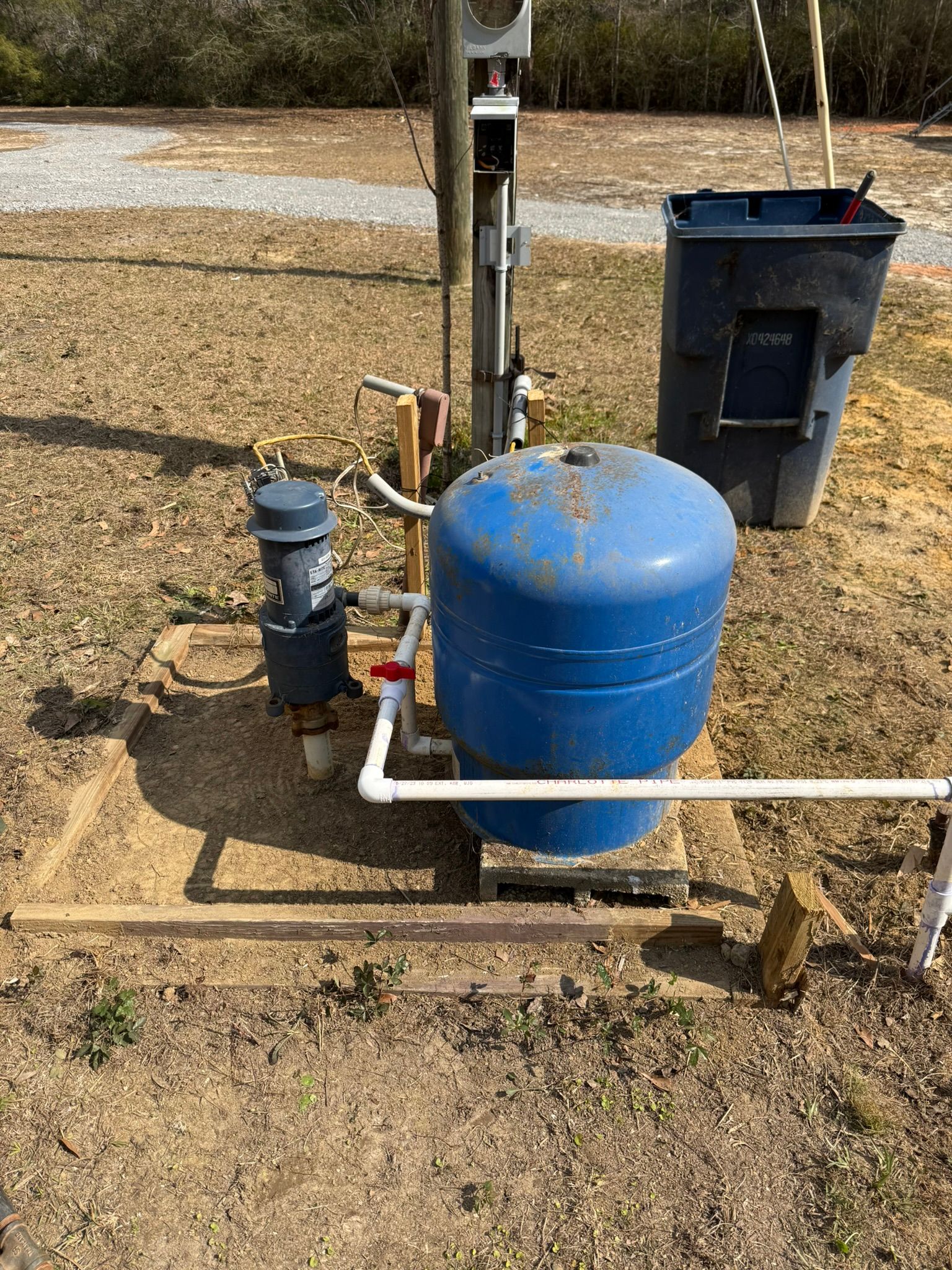 Blue pressure tank and plumbing equipment beside a black bin on a dirt patch outdoors.