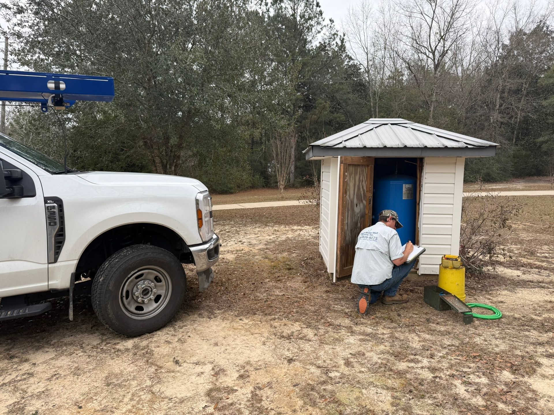 White utility truck beside a small shed; a worker kneels at the shed entrance with tools and a yellow bucket.