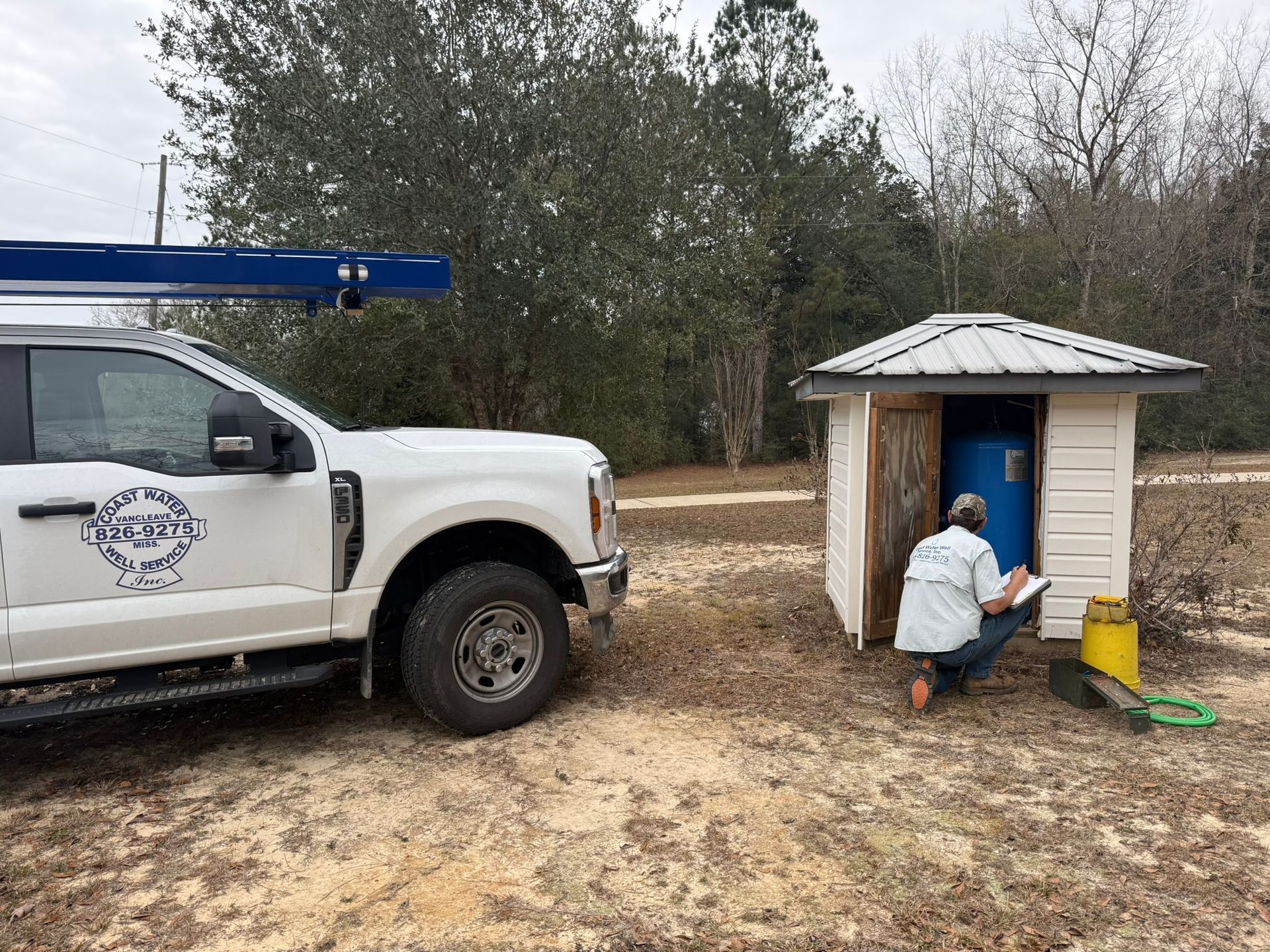 White utility truck beside a small shed; a worker kneels at the shed entrance with tools and a yellow bucket.