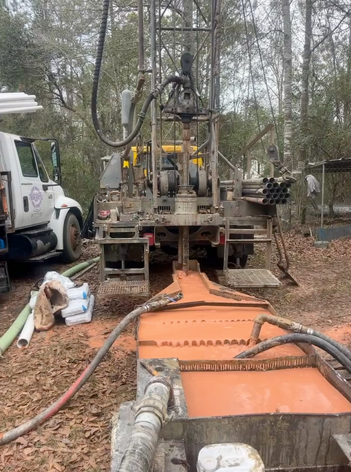 A drilling rig operates in a wooded area, discharging orange mud into a collection tank in the foreground.